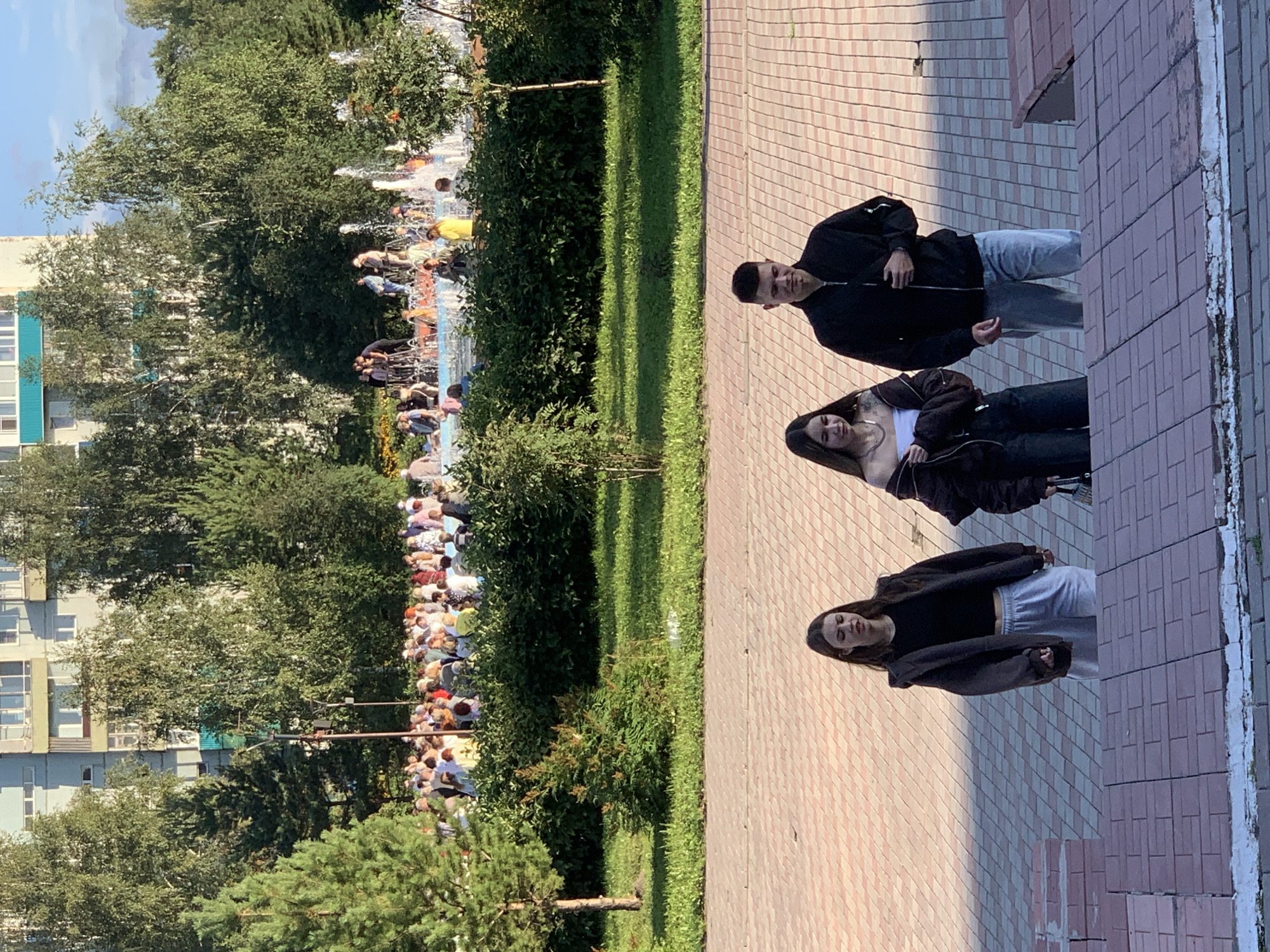An outdoor scene captures a group of young people walking down a paved stairway in Prokopyevsk, Russia. In the foreground, three individuals are visible: a woman on the left in a dark jacket and grey sweatpants, a woman in the middle wearing a dark jacket over a white top and black pants, and a man on the right in a black jacket and blue jeans. The stairway is made of brick and extends towards a patterned paved area. Behind them, a park setting unfolds with lush green grass, trees, and a large crowd gathered near a fountain. In the background, a multi-story building with windows is visible, along with a clear sky. The overall atmosphere suggests a warm, sunny day.