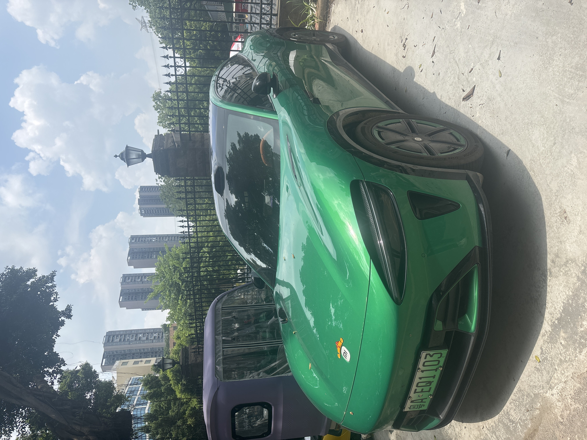 An exterior, eye-level, medium shot captures a vibrant green electric vehicle parked on a concrete surface. The car is positioned diagonally, facing towards the left of the frame, with its front end prominently displayed. The vehicle features sleek lines, a dark-colored roof, and distinctive headlights. A license plate with the characters "A-EQ1103" is visible on the front bumper. The car has a small, circular emblem on its hood with an orange figure. To the left of the green car, a portion of a purple three-wheeled vehicle is visible, covered with a clear plastic enclosure.
Behind the vehicles, a black metal fence runs horizontally, separating the parking area from a background scene. Beyond the fence, lush green trees and foliage are abundant. In the distance, several tall, modern residential buildings rise against a partly cloudy blue sky. A traditional lamppost is mounted on a stone pillar in the middle ground, further accentuating the blend of old and new in the urban landscape. The scene appears to be located in Nanhai District, China, as indicated by the context of the image's origin. The ground is littered with scattered dry leaves, suggesting an outdoor environment. The lighting indicates it is daytime.
