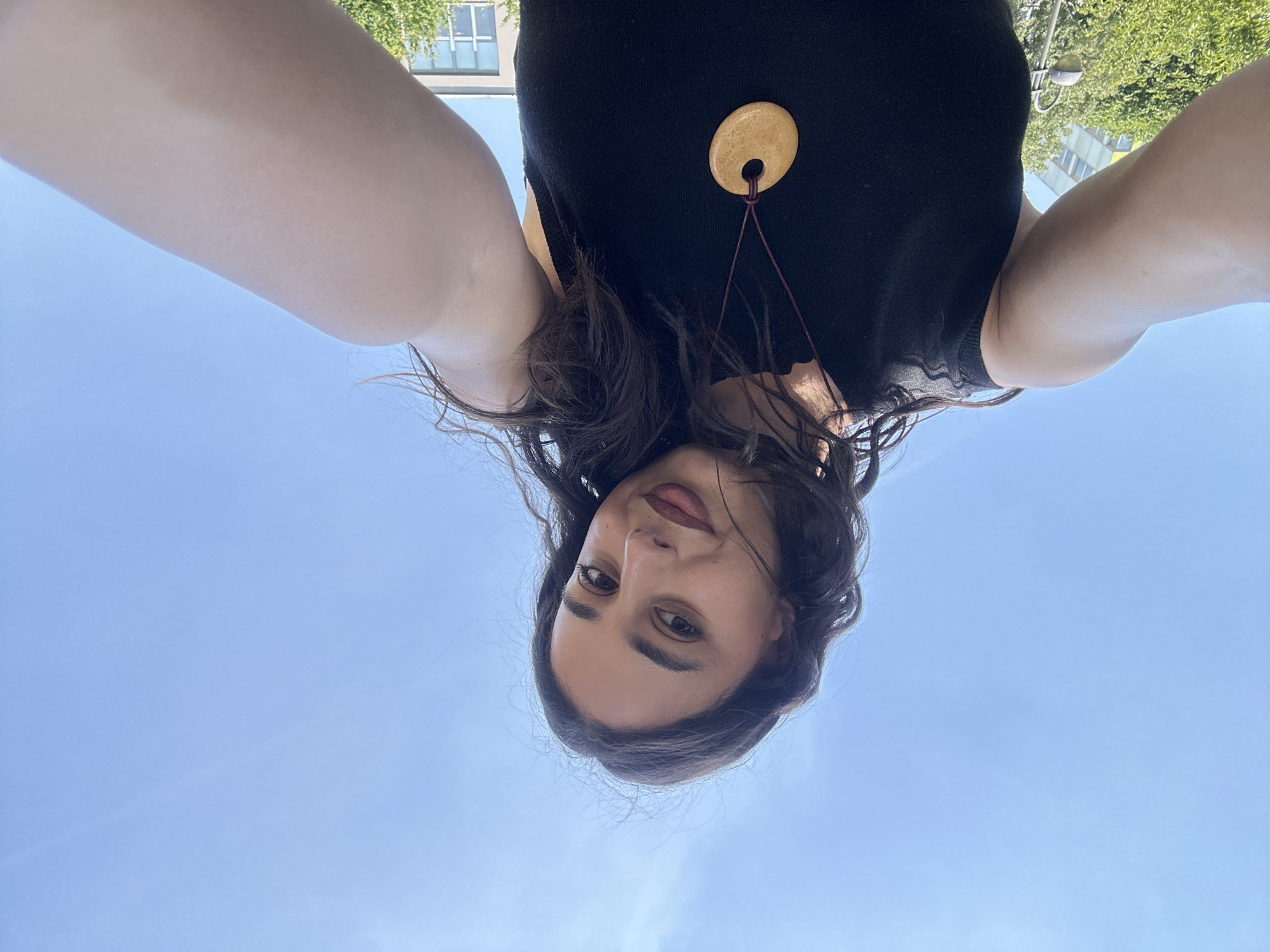 A low-angle, outdoor, selfie-style photograph shows a young woman with dark, wavy hair and dark eyes against a bright blue sky. She is wearing a black sleeveless top and a light-colored pendant on a cord necklace. Her right arm is extended, holding the camera, with her bicep and shoulder in focus. In the background, out-of-focus elements include green trees, a lamppost, and buildings. The bright sky and sun suggest a daytime setting. The woman's expression is a slight smile.