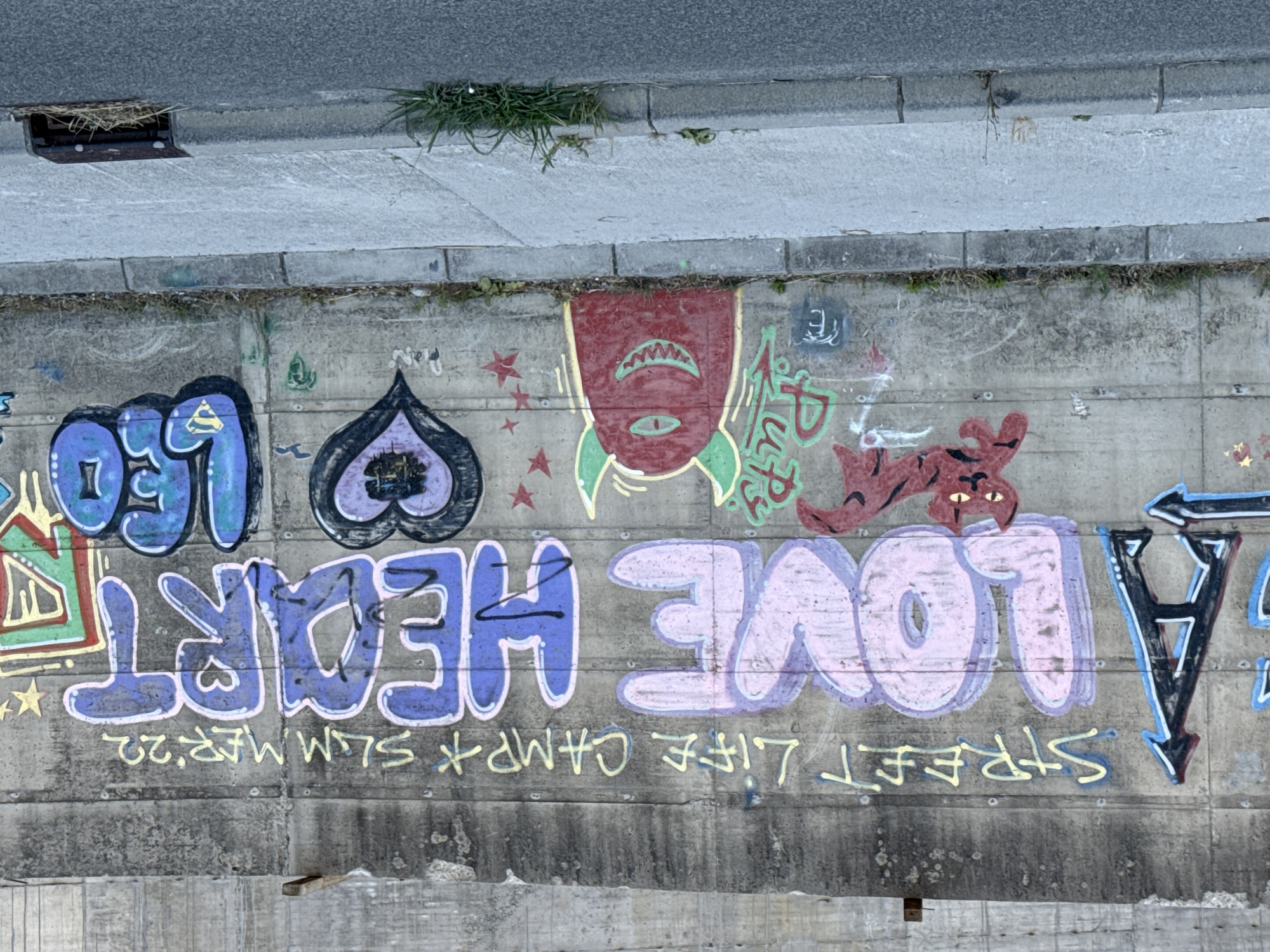 A wide-angle, eye-level shot captures a section of a concrete wall covered in graffiti, with a sidewalk and road in the foreground. The graffiti features large, colorful lettering. Dominating the center are the words "LOVE HEART" in large, bubble-style letters, with "LOVE" in pink and "HEART" in blue. To the left of "LOVE," a large black and blue letter "A" with an arrow pointing left is visible. To the right of "HEART," the word "LEO" is written in large blue letters, with a yellow Superman emblem on the "E." Above the main lettering, the words "STREET LIFE CAMP SUMMER '22" are scrawled in yellow. Various smaller graffiti elements are present, including a red cat with a black outline, a red cyclops monster with green horns and a wide grin, and a black heart with purple and black outlines. The concrete wall shows signs of weathering and stains. The sidewalk is gray concrete, with some patchy grass and debris. A storm drain grate is visible at the edge of the road. The scene appears to be in an urban or semi-urban environment, likely a public space. There are no people visible in the image. The location is identified as Boreti, Montenegro.