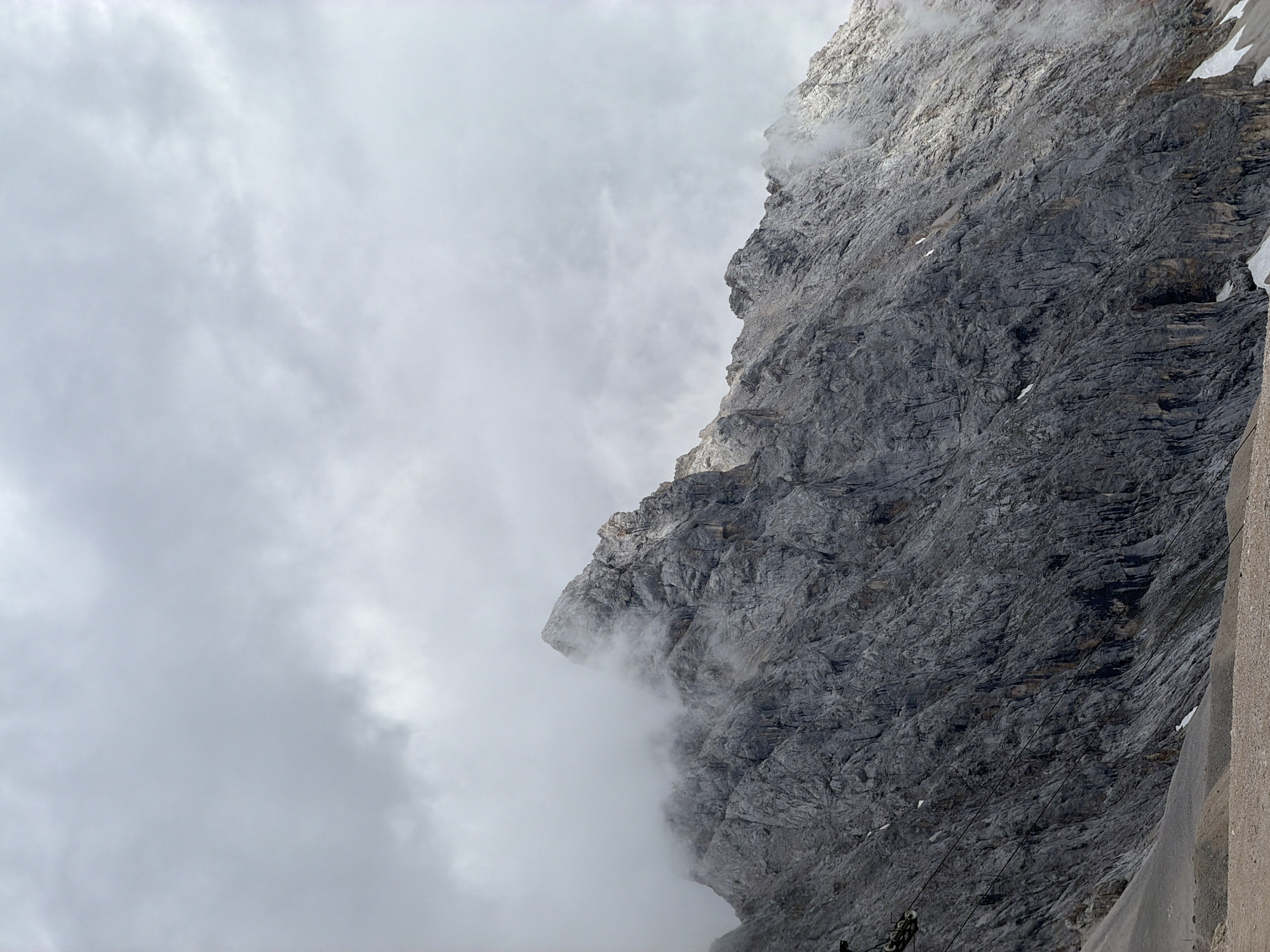 The image shows a dramatic, close-up view of a steep, rocky mountain face. The rock is heavily textured and appears weathered, with layers and fissures visible. Clouds or mist are partially obscuring the upper and left portions of the mountain, creating a sense of atmospheric depth and mystery. The lighting suggests it could be daytime, but the overcast conditions and mist create a muted and somewhat somber mood.
On the right side of the image, a light-colored, smooth, and likely man-made structure runs vertically, contrasting with the rugged natural texture of the mountain. Thin, dark lines, possibly cables, stretch across the rock face in some areas, suggesting some form of infrastructure or activity related to the mountain, such as a cable car system. A small amount of snow is visible on some ledges of the mountain, hinting at the high altitude and potentially cold weather.
The scene is devoid of people, focusing entirely on the imposing natural landscape and the interaction with man-made elements. The overall impression is one of a raw, powerful natural environment, potentially a challenging or awe-inspiring location. Given the location context of Garmisch-Partenkirchen, Germany, this could be a view of the Zugspitze or a similar prominent Alpine peak. The weather appears to be cloudy and possibly a bit windy, given the way the mist is swirling around the mountain.