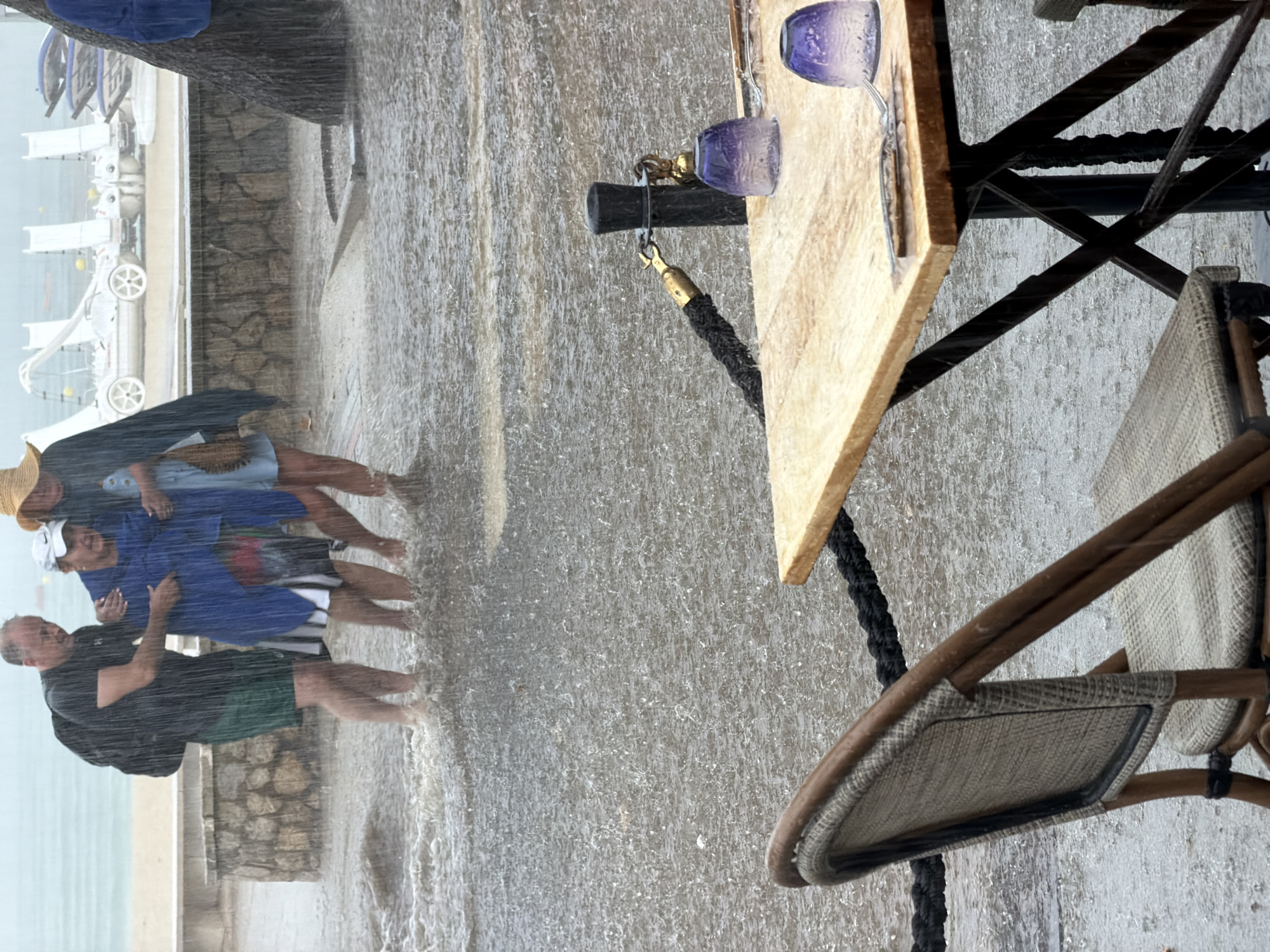 In Palmanova, Spain, amidst a downpour, three individuals are wading through ankle-deep water on a paved area near a beach. A man on the left, wearing a dark t-shirt and green shorts, carries a black backpack and supports a woman in the center. The woman is wrapped in a blue blanket and appears distressed. To her right, another woman in a light blue dress and a straw hat is being aided. In the foreground, an empty outdoor cafe table is set with a fork and knife, alongside two purple glasses, one of which is partially filled with water. A wicker chair is positioned at the table. Black ropes, possibly marking a boundary, are visible. The background shows a stone wall, beach umbrellas, and the sea.