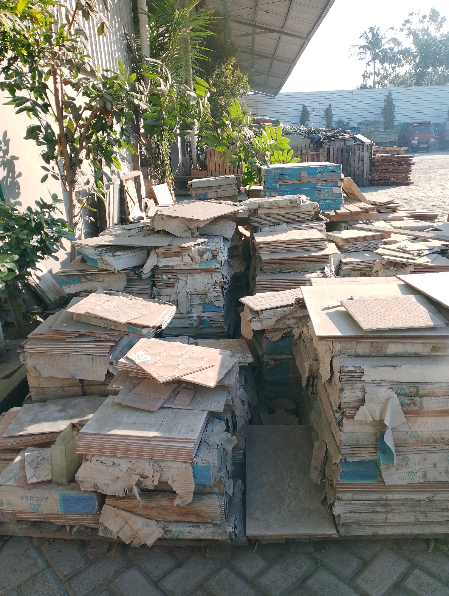 In Jambean, Indonesia, an outdoor storage area is filled with stacks of tiles, many of which are damaged or discarded. The tiles are of various sizes and patterns, some with hexagonal designs, others with geometric or plain surfaces. They are piled high on wooden pallets and directly on the paved ground. Cardboard packaging, some torn and weathered, is interspersed with the tiles. In the background, a building with a corrugated metal roof is visible, along with lush green foliage from trees and plants. Further back, more piles of materials, including wooden planks and possibly construction debris, are present. A red truck is parked in the distance. The scene appears to be a commercial or industrial yard, likely a tile supplier or a construction waste disposal site.