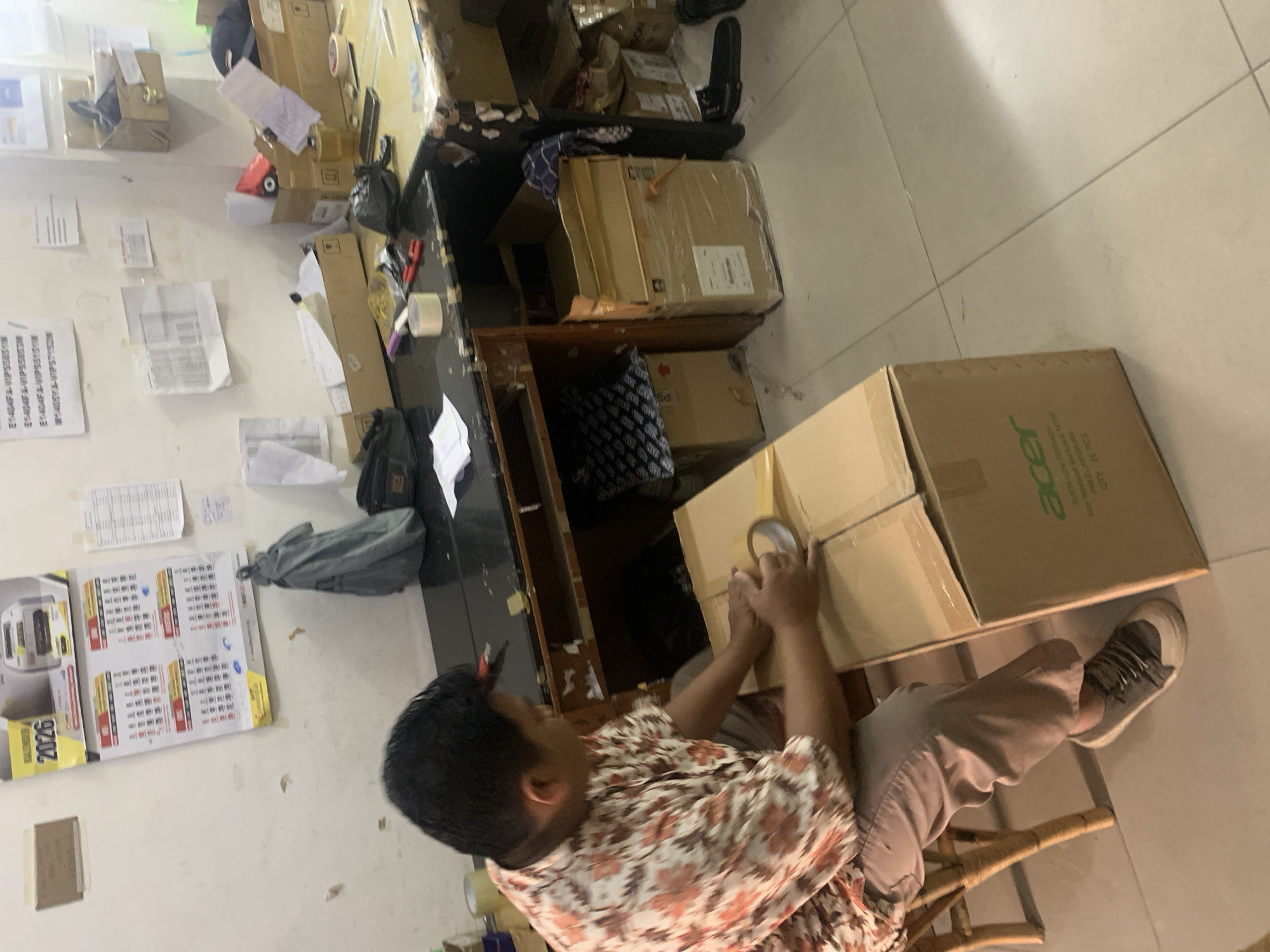 The image depicts an indoor scene, likely an office or storage area, within Palembang, Indonesia. A man, wearing a floral-patterned shirt and light-colored pants, is seated on a stool, actively taping a cardboard box. The box is positioned on the floor in front of him. The environment appears cluttered, with various items scattered on shelves and the floor, including other cardboard boxes, papers, and office supplies. A calendar is visible on the wall, displaying the year 2026. The overall impression is of a workspace where packaging or shipping activities are taking place.