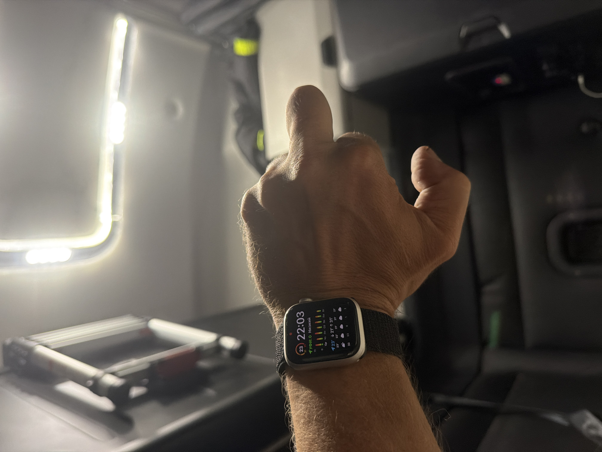 An individual's left wrist, adorned with a dark fabric watch band and a smartwatch, is positioned in the foreground. The smartwatch displays the time as 22:03 and features various weather-related icons and data. The background suggests the interior of a vehicle, possibly a truck cab, with a bright light source overhead and dark upholstery visible. The scene appears to be dimly lit, indicating a nighttime or low-light environment. There are no visible landmarks or indicators to place this image in New Britain, United States of America.