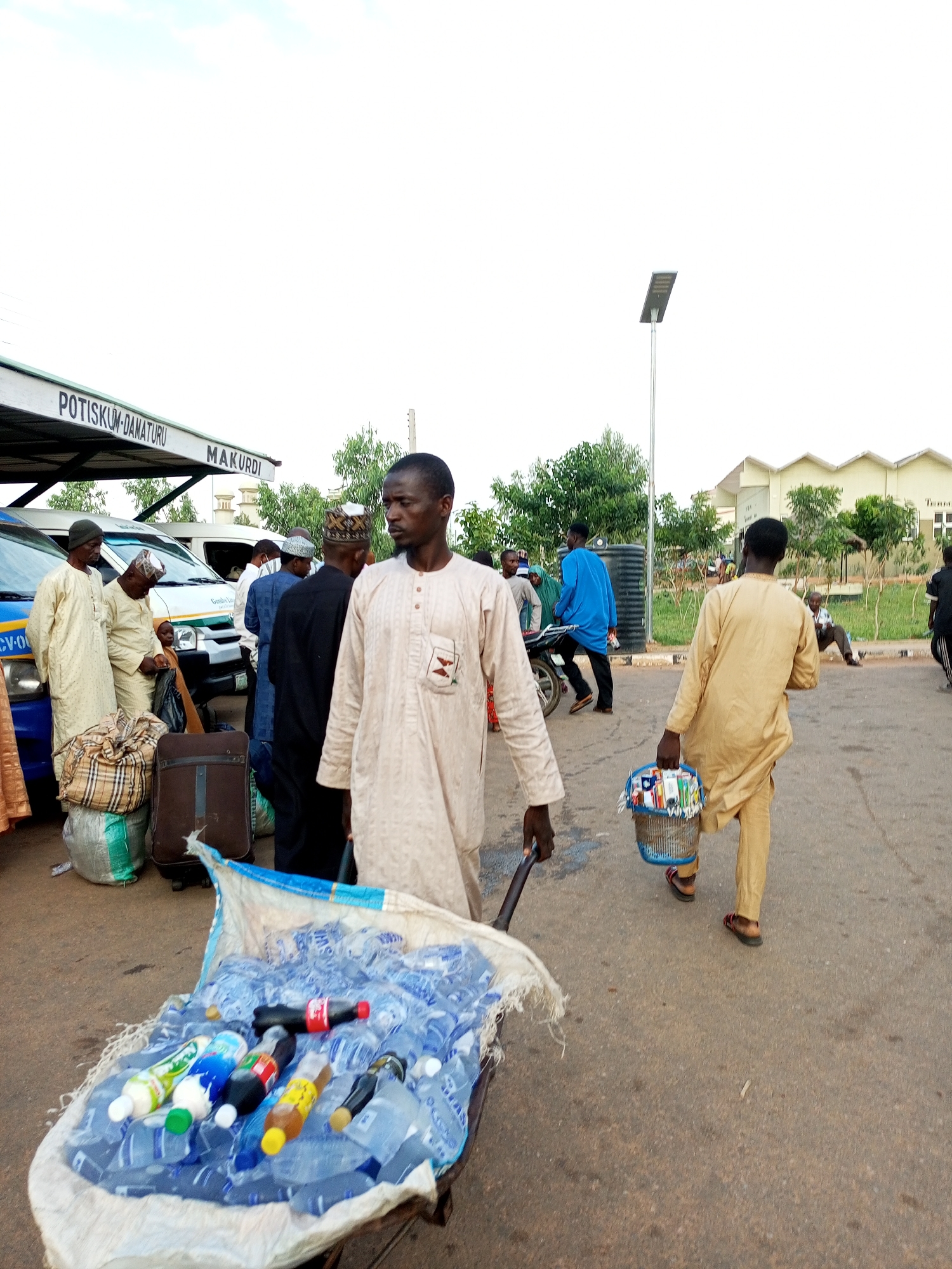 An outdoor medium shot shows a scene at a bus station in Gombe, Nigeria. The sky is overcast with a few wispy clouds. In the foreground, a man in a light brown robe pushes a cart filled with bottled drinks. The cart is draped with a blue and white tarp. Behind him, another man in a similar robe walks away from the camera, carrying a basket of items. To the left, a group of men dressed in traditional attire stand near a blue and white bus. Luggage is piled on the ground. In the background, a modern building with a beige, segmented facade is visible, along with trees and a lamppost. The ground is paved.