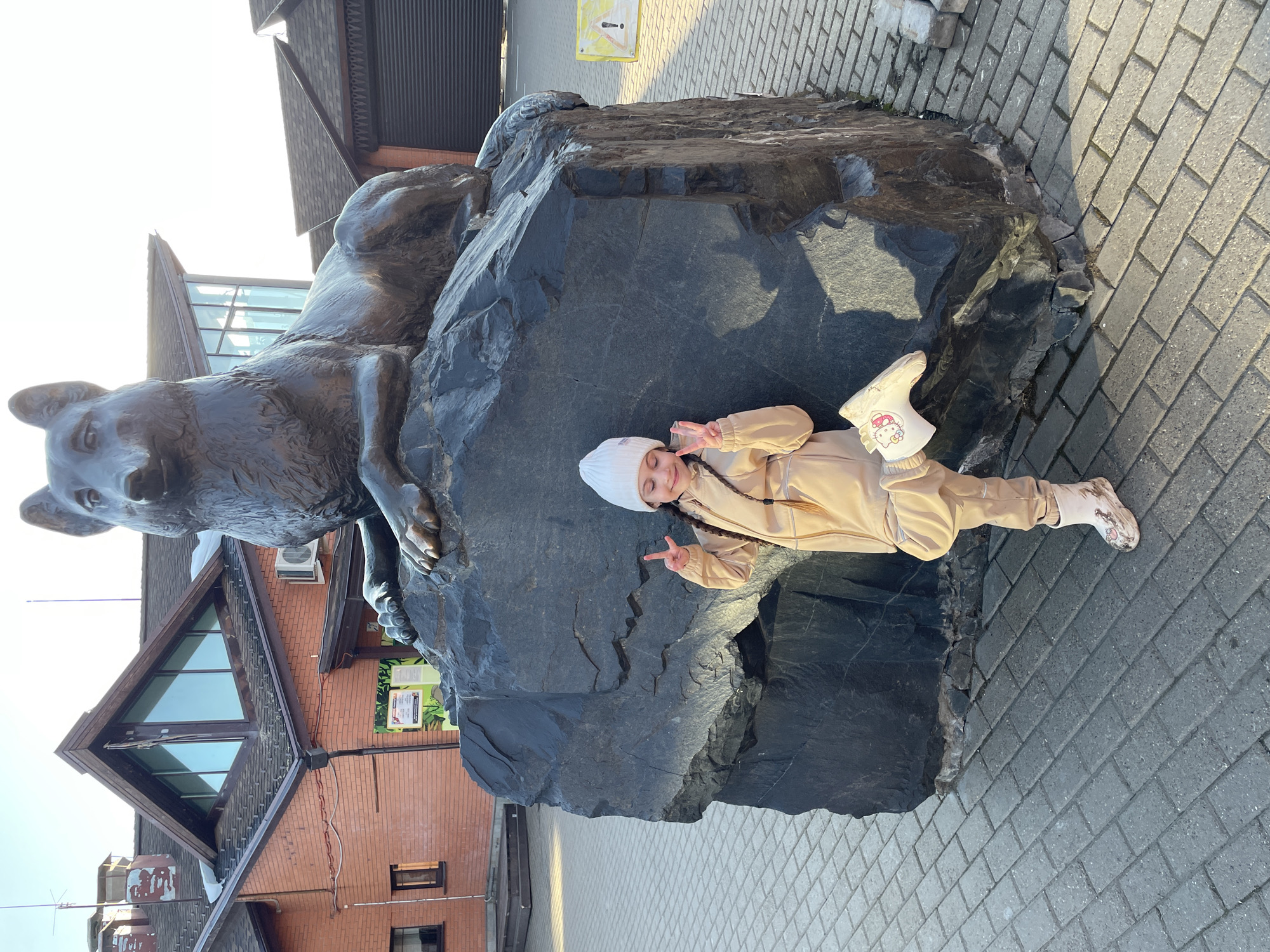 A young girl poses next to a dark gray stone sculpture of a wolf located on a paved brick surface in Izhevsk, Russia. The sculpture, situated on a large rock, has an air conditioning unit and informational sign nearby. The girl, wearing a beige outfit and white hat and boots, is lying on the rock and holding up two fingers in a peace sign. A red brick building with a unique peaked roof stands in the background.