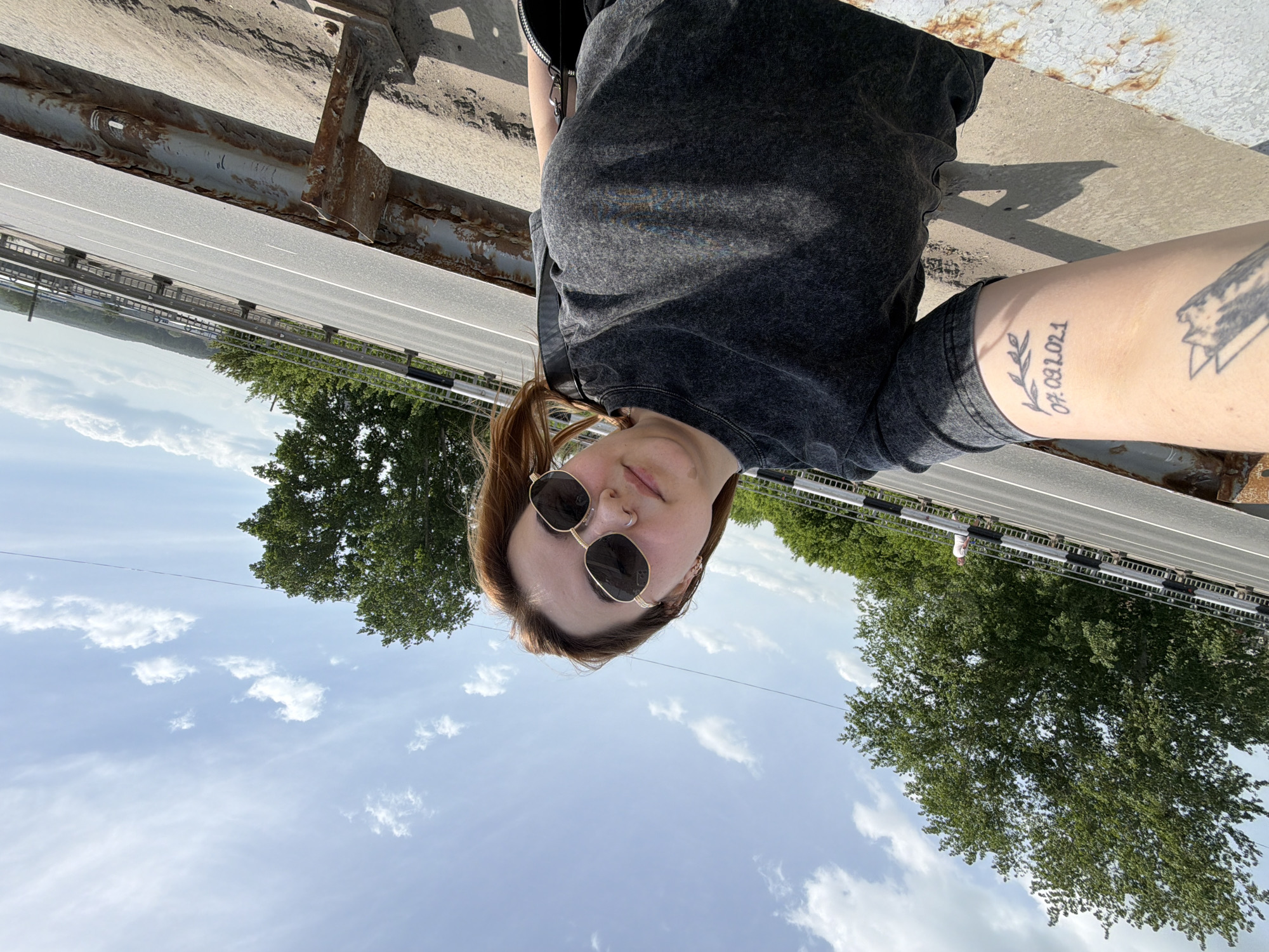 A selfie-style photograph shows a person standing on a bridge or elevated walkway in Chaikovsky, Russia, as indicated by the surroundings visible in the background. The person has light skin, round sunglasses, and their hair is pulled back in a ponytail. They are wearing a dark-colored t-shirt. The individual's left arm is visible, displaying two tattoos on the inner forearm, one with a date and leaf design and another of a symbolic nature. The scene includes a view of a highway, trees, and a sky with clouds. The bridge or walkway has a weathered metal railing.