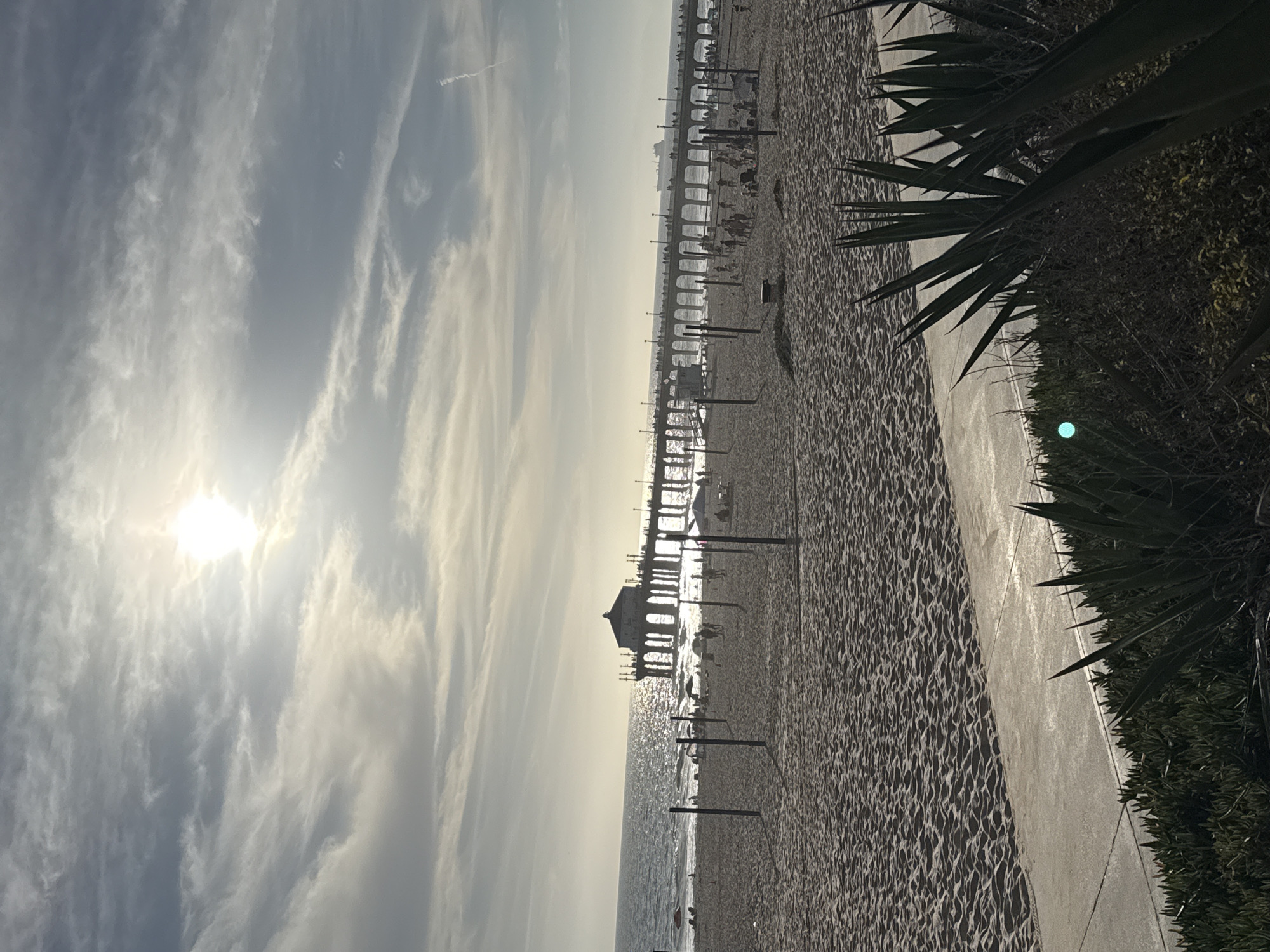 The image captures a wide shot of the Manhattan Beach Pier in Manhattan Beach, California, USA. The pier extends into the ocean, with its distinctive octagonal building visible at the beginning of the pier structure. Numerous people are seen walking along the pier, and several boats are visible in the distance. The beach in the foreground is sandy, with volleyball nets set up. The ocean waves are gently rolling onto the shore. The sky is partly cloudy, with the sun partially obscured by clouds, casting a soft light on the scene. The overall atmosphere is serene and depicts a typical day at the beach with people enjoying recreational activities.