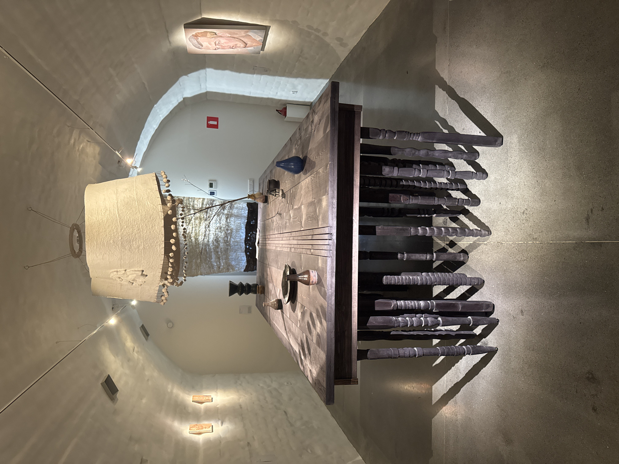 The image shows an art installation in a vaulted, white-walled room, possibly a museum or gallery. The room is lit by spotlights and overhead fixtures. The central focus is a large, dark wooden dining table with ornately carved legs. The table is set with several ceramic vases and a plate. Above the table hangs a large, textured lampshade adorned with small round objects.
On the left wall, there are two small, framed artworks. On the right wall, a framed portrait of a man is displayed. The floor is concrete.
While the image depicts an art installation, it does not provide any specific details that would place it in Suzdal, Russia, or any other specific city. The environment appears to be a generic exhibition space.