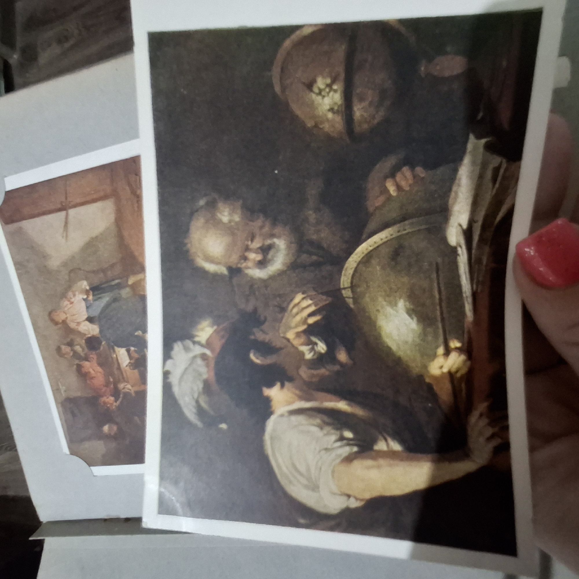 The image displays two paintings, one on top of the other. The top painting, partially visible and framed, depicts a group of figures gathered around a table in what appears to be an indoor setting with natural light. The bottom painting, in the foreground, is a close-up view of two men and several globes. One man, with a long white beard, looks intently at a globe in front of him, while a younger man, wearing a light-colored shirt, leans in to examine it as well, pointing with a tool. Another globe is visible to the right. The overall tone of the bottom painting is dark and atmospheric. The context of the location in the city of Unknown, Unknown is not discernible from the image.