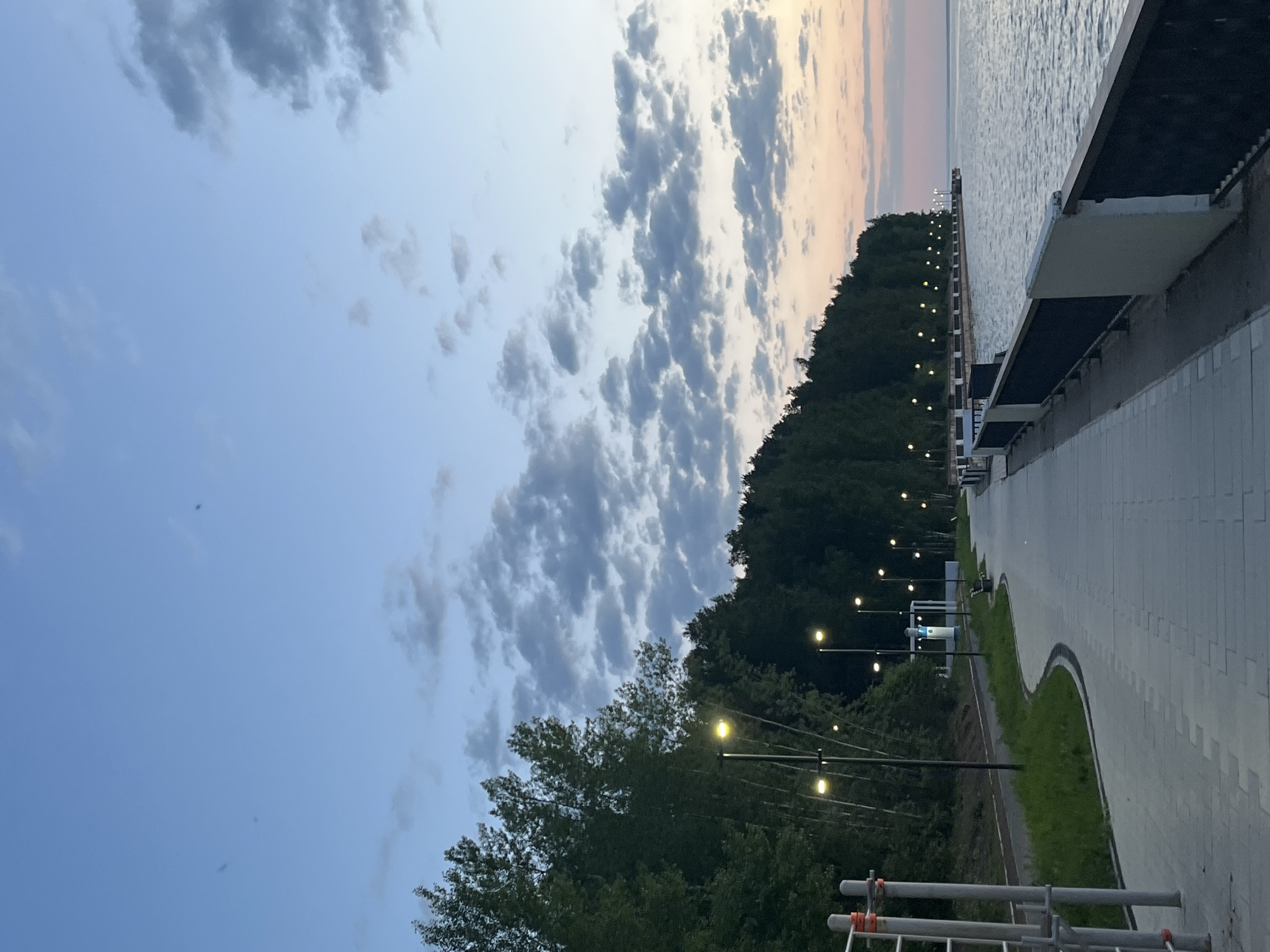 The image shows a waterfront promenade in Chaikovsky, Russia, at dusk. The sky transitions from blue to a pale orange above the water. The scene includes a paved walkway lined with trees and evenly spaced streetlights illuminating the path. A modern building structure extends along the waterfront. There are few people visible. The image has been rotated 90 degrees counterclockwise.