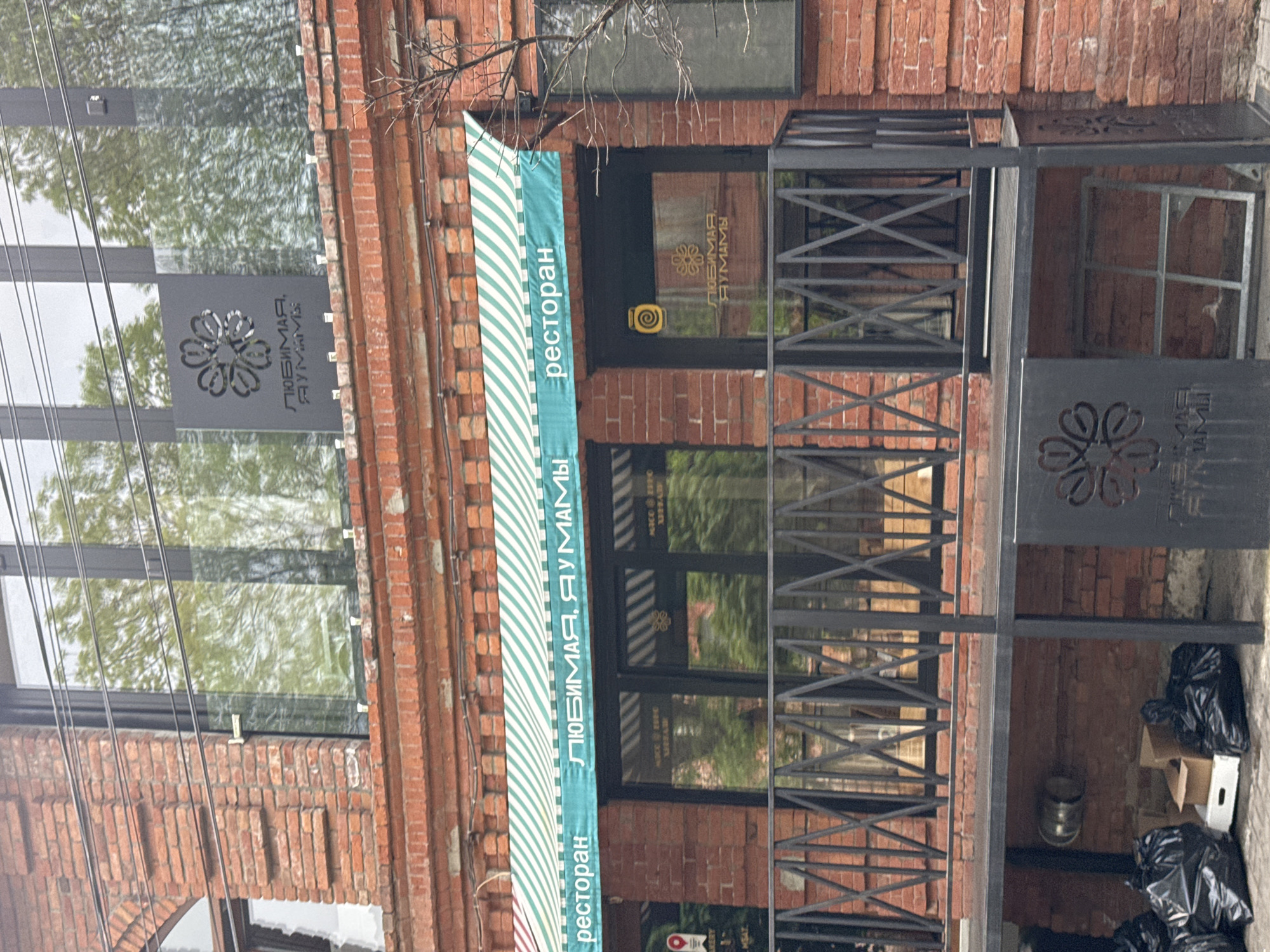 The image depicts the exterior of a restaurant called "Lyubimaya, Dumami" ("Beloved, Dumami") located in Akhieli, Georgia. The restaurant is housed in a brick building with a striped awning. The signage includes the name of the restaurant in Russian, accompanied by a floral logo. A black metal structure, possibly a decorative railing or small porch, is present in front of the entrance. Trash bags and cardboard boxes are visible near the base of the structure. Power lines run along the top of the building.
