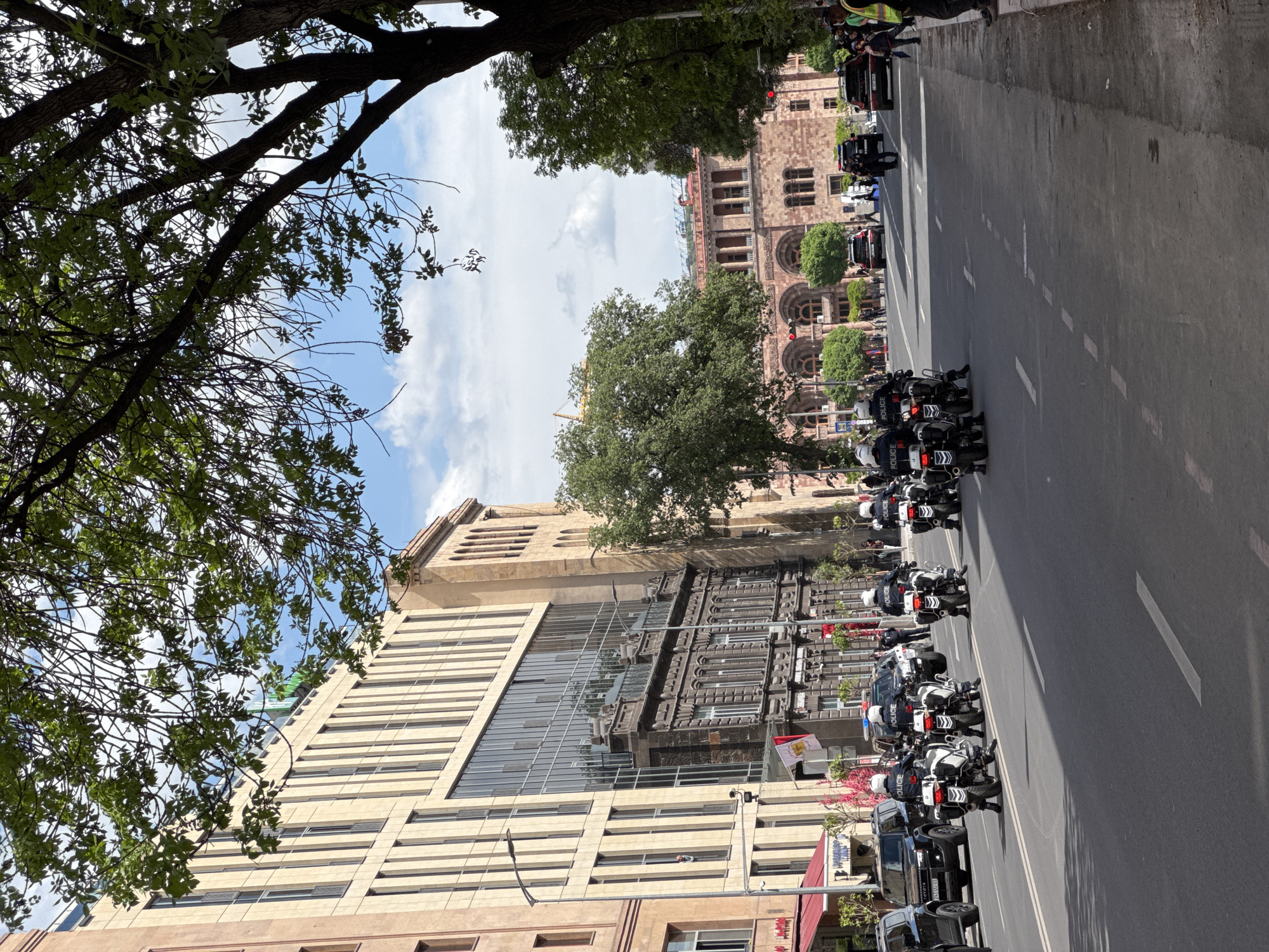 A Yerevan, Armenia city street scene shows a motorcade of police motorcycles and black SUVs driving on a city street. Trees line the street on both sides of the road. Buildings are on both sides of the street, including one with a modern, glass-fronted section. The sky is blue with white clouds. People are visible on the sidewalks. A flag of Egypt is visible on a pole.