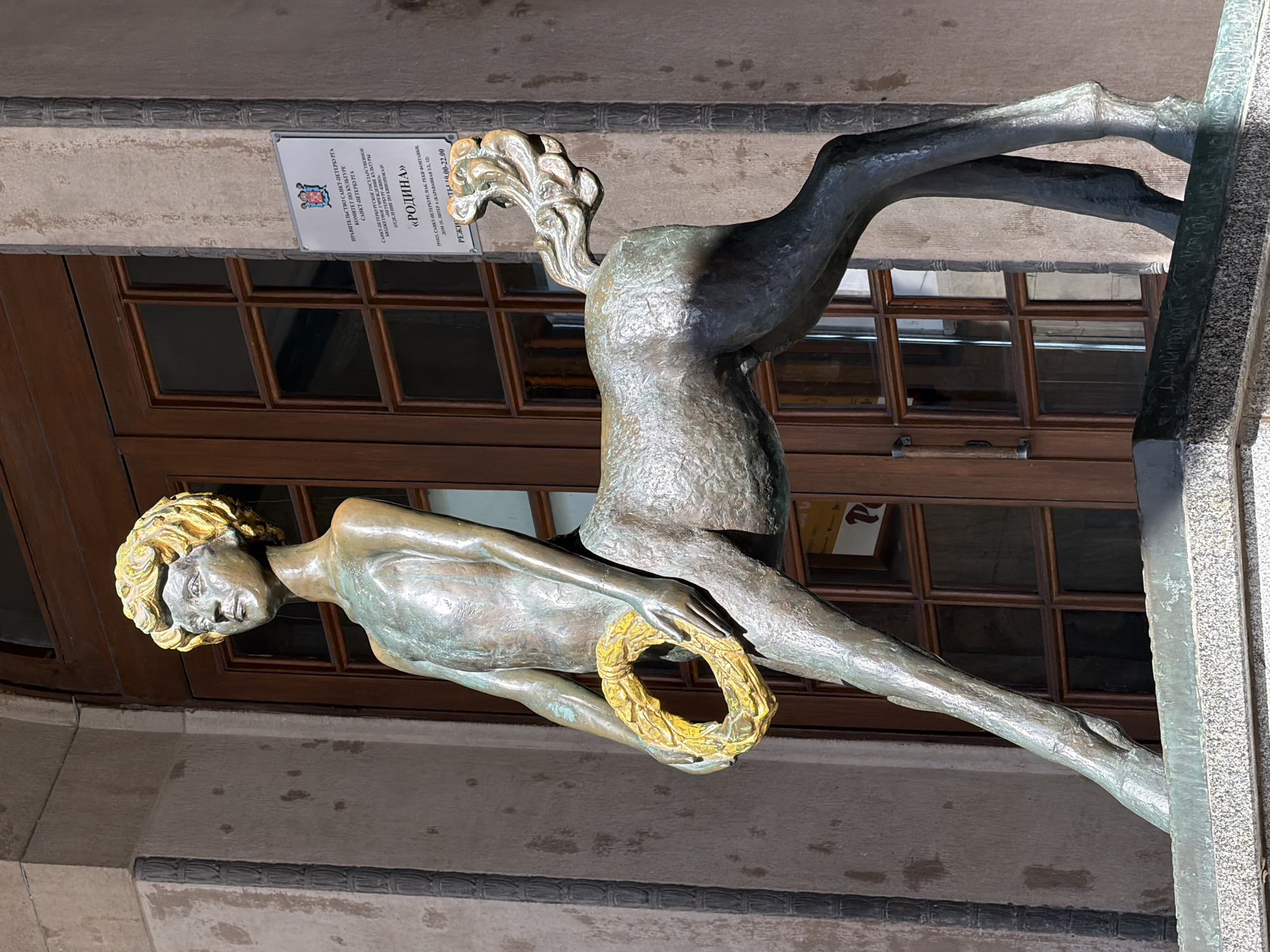 An ornate bronze sculpture, photographed on its side, is the central subject, depicting two intertwined or overlapping human-like figures. The lower figure, appearing to be a man, lies reclined with his upper body visible, holding a golden wreath in his hands. He has distinctive golden, wavy hair and a contemplative expression. Above him and behind his back, another stylized figure, possibly abstract or female, extends upwards and to the right, characterized by a detailed, organic, wavy form where a head might be.
The sculpture is positioned in front of a building facade with dark wooden-framed windows, reflecting some interior light. The background elements suggest an urban outdoor setting during daylight, with bright light reflecting off the bronze, highlighting its textures and the gold accents.
A white plaque affixed to the wall above the sculpture provides significant location context. Visible Russian text on the plaque identifies the entity as the "Government of St. Petersburg, Committee for Culture of St. Petersburg," and specifically mentions the "St. Petersburg State Budgetary Cultural Institution" and the "Cinema Exhibition Department" named "Rodina" (Homeland/Motherland). An address is also provided: "191023, St. Petersburg, Fontanka River Embankment, House 15, Letter A (Karavannaya St., 12)." There's also a mention of "РЕЖИМ" (Mode/Hours) and "19.00-22.00", likely indicating operational hours for the cinema. The location is confirmed as Saint Petersburg, Russia, based on the text.