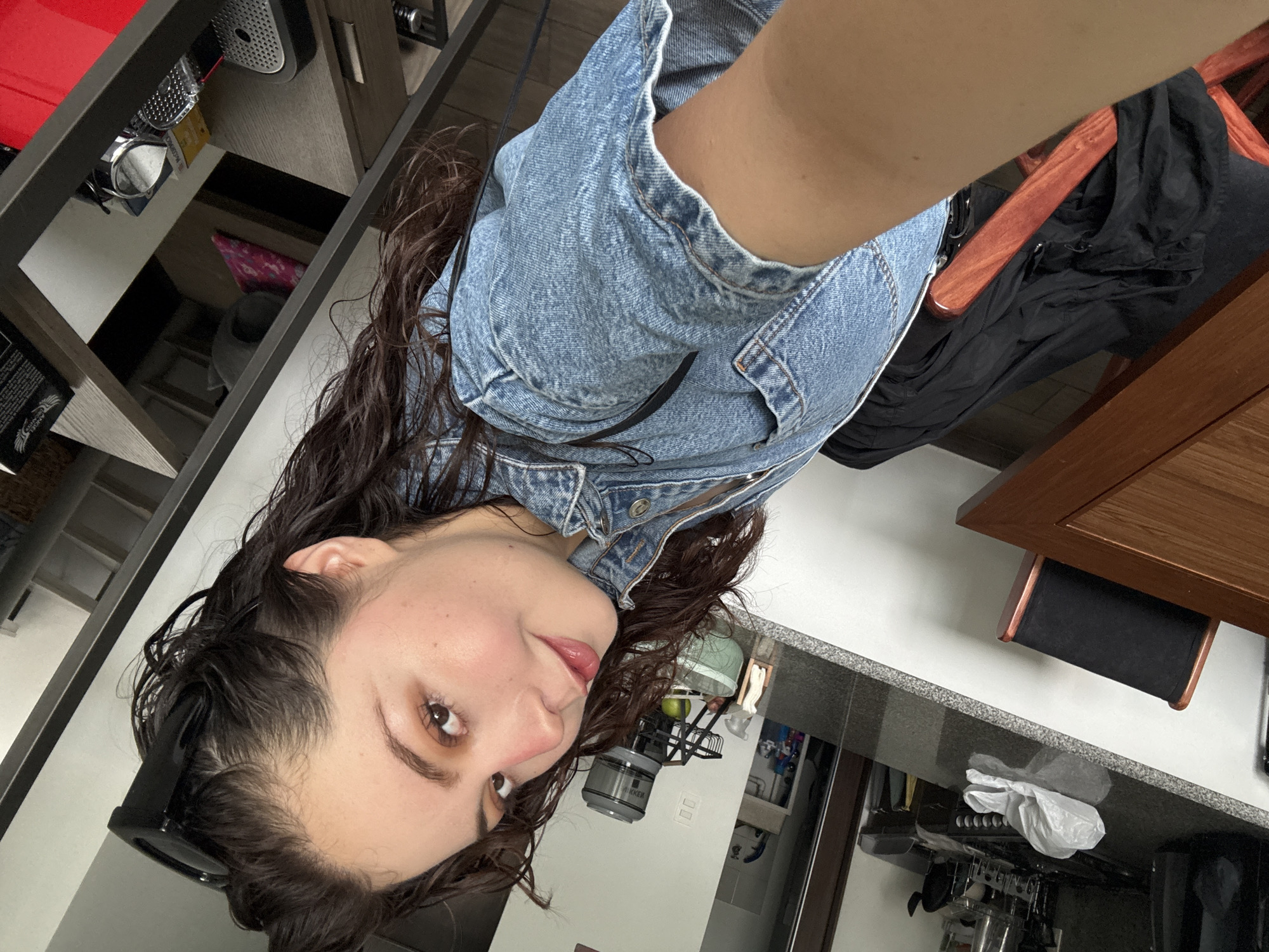 The image is a selfie taken indoors, likely in a kitchen. A person with long, dark, wet hair is wearing a denim jacket. They are looking at the camera. The background reveals a kitchen counter with various objects including a blender, dish rack, and kitchen utensils. Part of a wooden table and chair is visible. The location is inferred to be in Mexico City, Mexico, based on potential cultural or architectural cues within the kitchen's design and items, though no definitive markers are visible.
