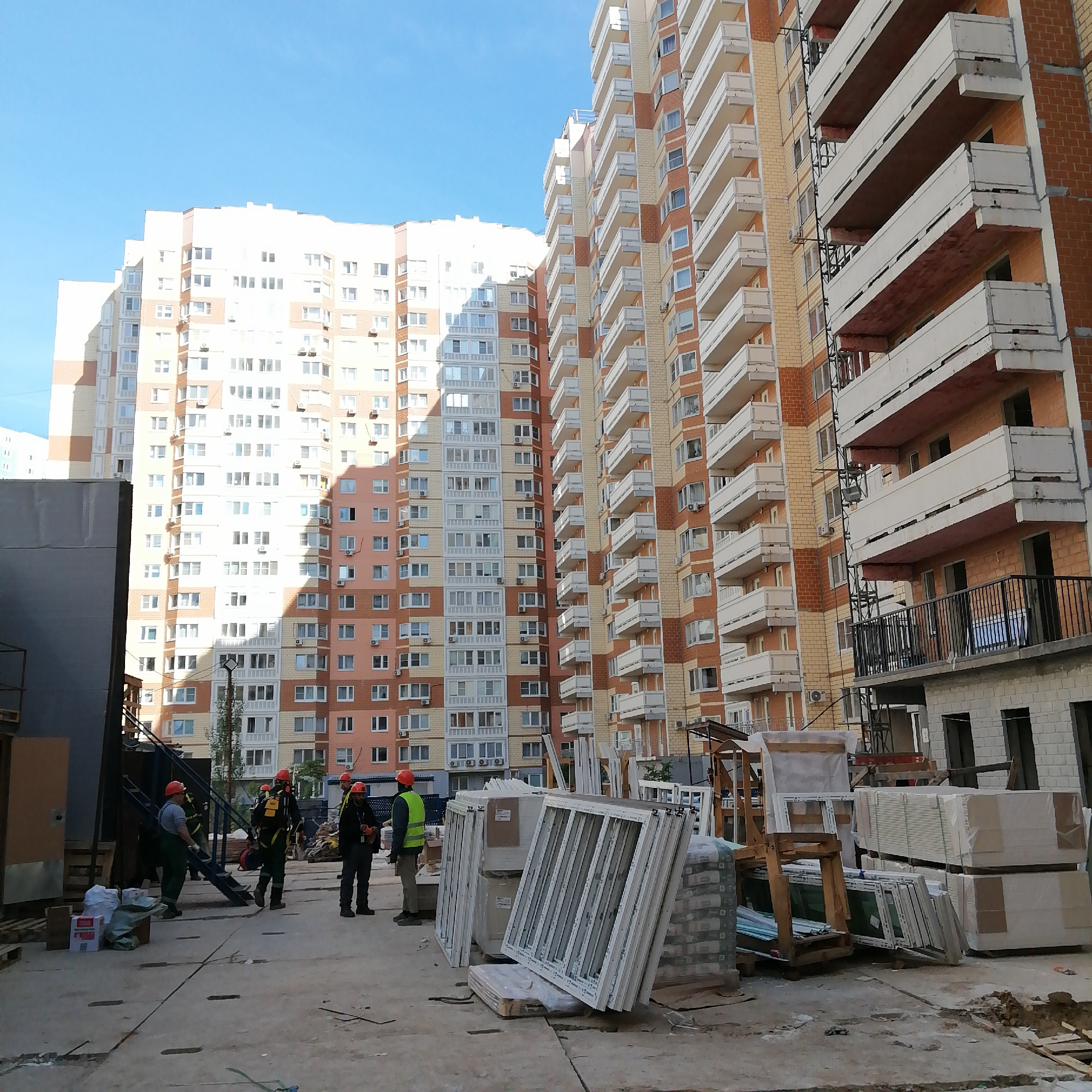 The image shows a construction site located in an urban area. Several construction workers wearing hard hats and work clothes are present. There are stacks of windows, building materials, and construction equipment on the concrete ground. In the background are large, multi-story apartment buildings with balconies. The sky is clear and blue. The overall scene suggests ongoing construction work in a residential area.