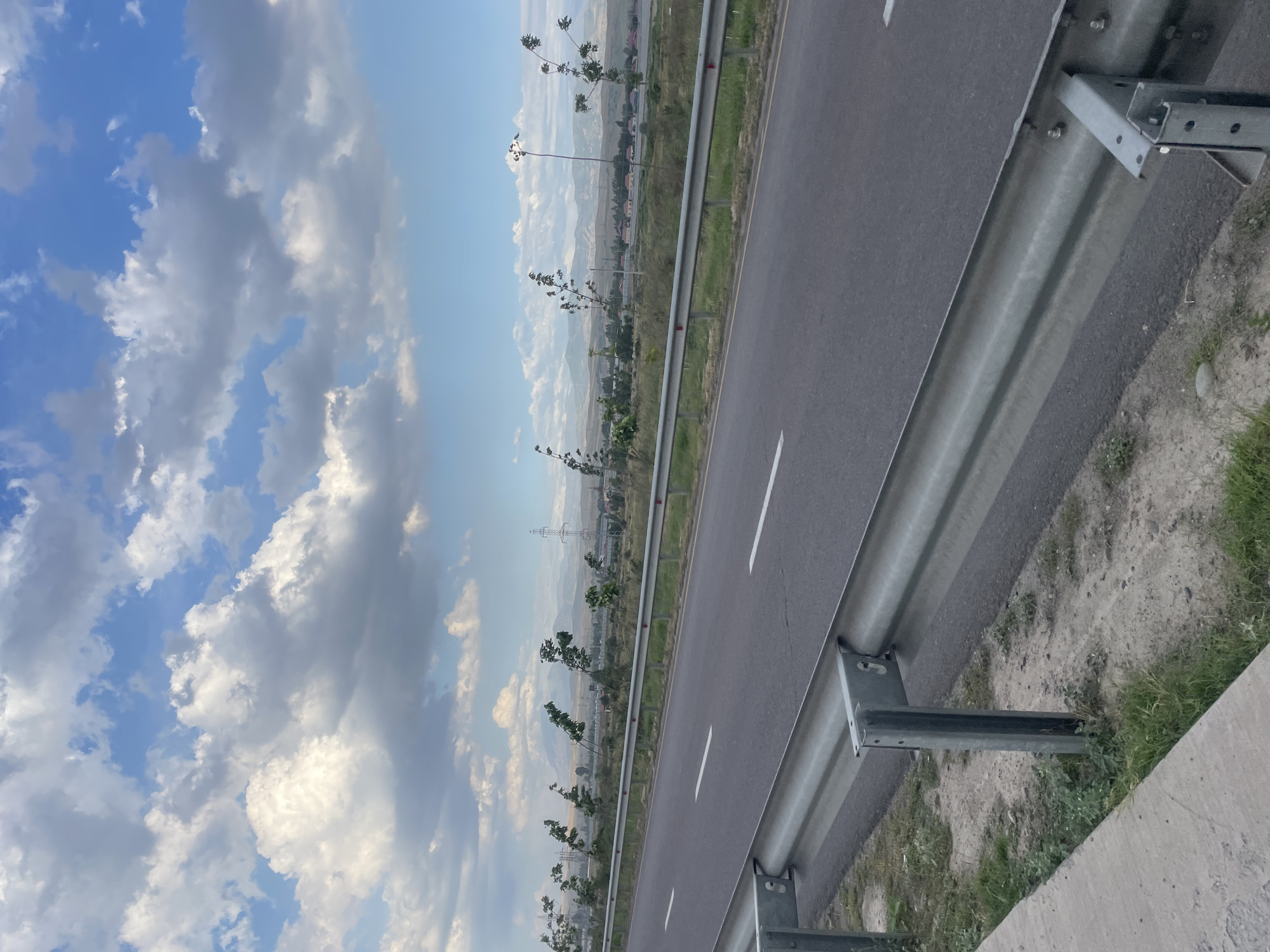 The image depicts a highway with a guardrail in the foreground. The road is asphalt with white lane markings. Beyond the guardrail is a grassy area with sparse vegetation. In the distance, there are trees, power lines, and mountains under a cloudy sky. The scene appears to be an open area with no visible people or buildings in the immediate vicinity. The lighting suggests it is daytime.