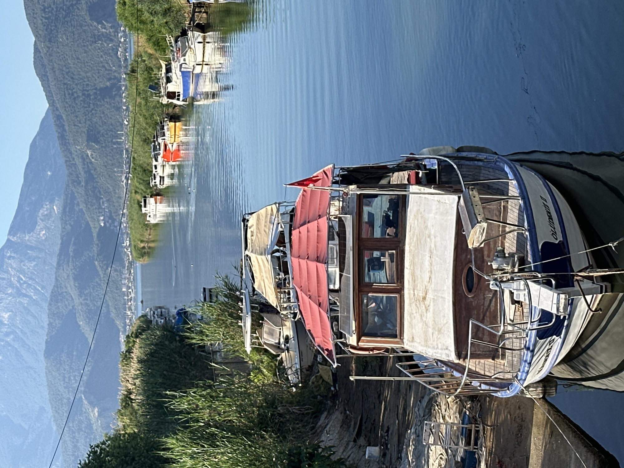 The image depicts a waterway lined with boats in Fethiye, Turkey. In the foreground, a large boat with a wooden cabin and a red awning is docked. Other boats of varying sizes and colors are moored along the waterway. The background features a mountain range and a glimpse of the city. Lush vegetation lines the shore near the boats. The water is calm and reflects the sky and surrounding landscape.