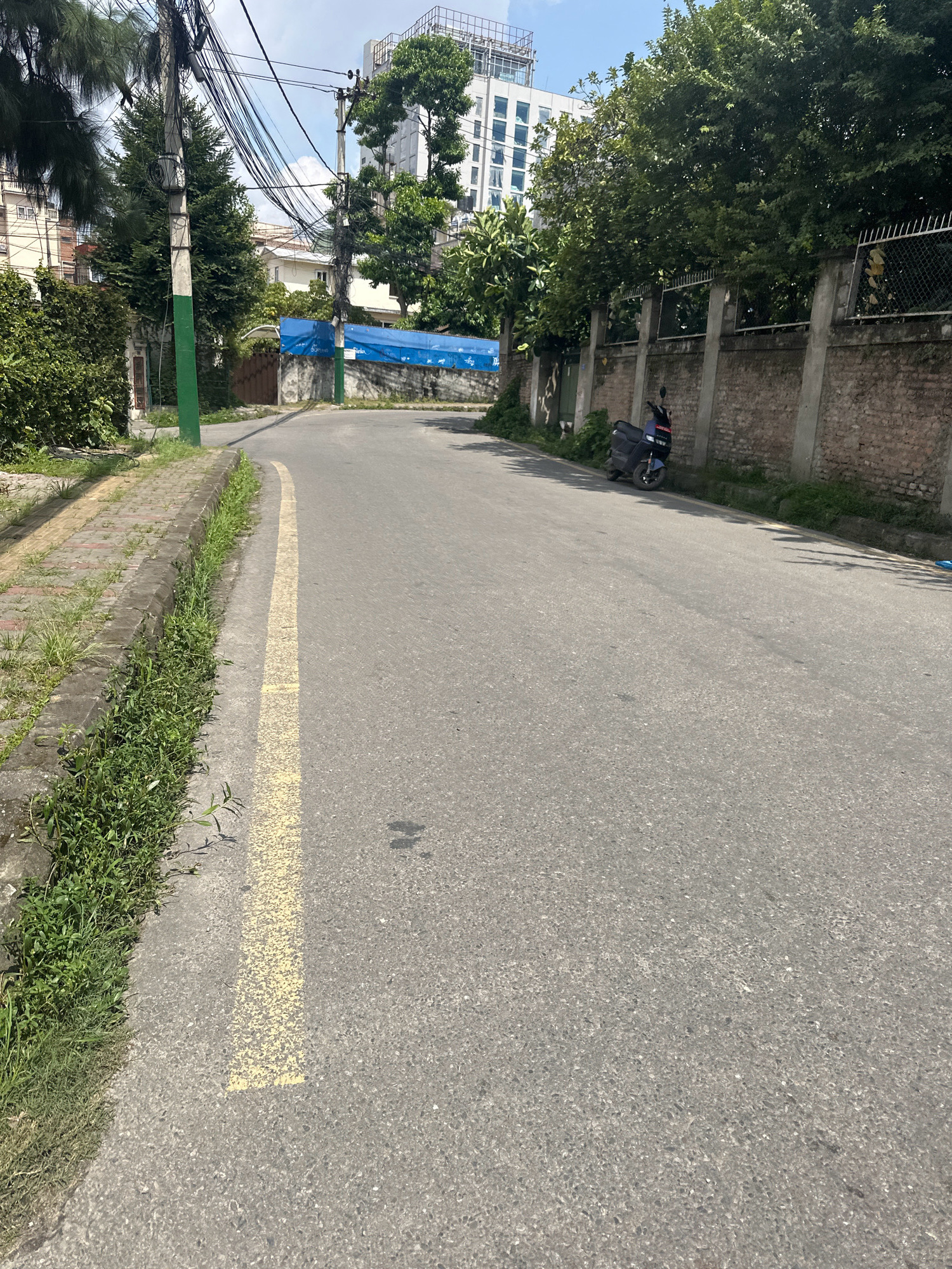 The image depicts a street scene in Lalitpur, Nepal, on a clear day. The road, paved with asphalt, features a yellow line marking a lane. To the left of the road is a sidewalk constructed with interlocking concrete pavers, bordered by a curb with overgrown grass and vegetation. Beyond the sidewalk, there are various elements of urban infrastructure including utility poles with a dense network of electrical wires and trees.
Further down the road, a dark blue scooter is parked along the right side. Adjacent to the scooter is a high brick wall with concrete pillars and a metal fence on top, separating the street from the property behind it. Behind the wall, lush green trees and some buildings are visible, including a tall white building with multiple floors and rows of windows. A large blue tarp is stretched across an area behind a section of the wall, possibly covering something outdoors. The environment appears to be a mix of residential and commercial or institutional development, typical of a city setting. The lighting suggests it is daytime, with shadows indicating the sun's position.