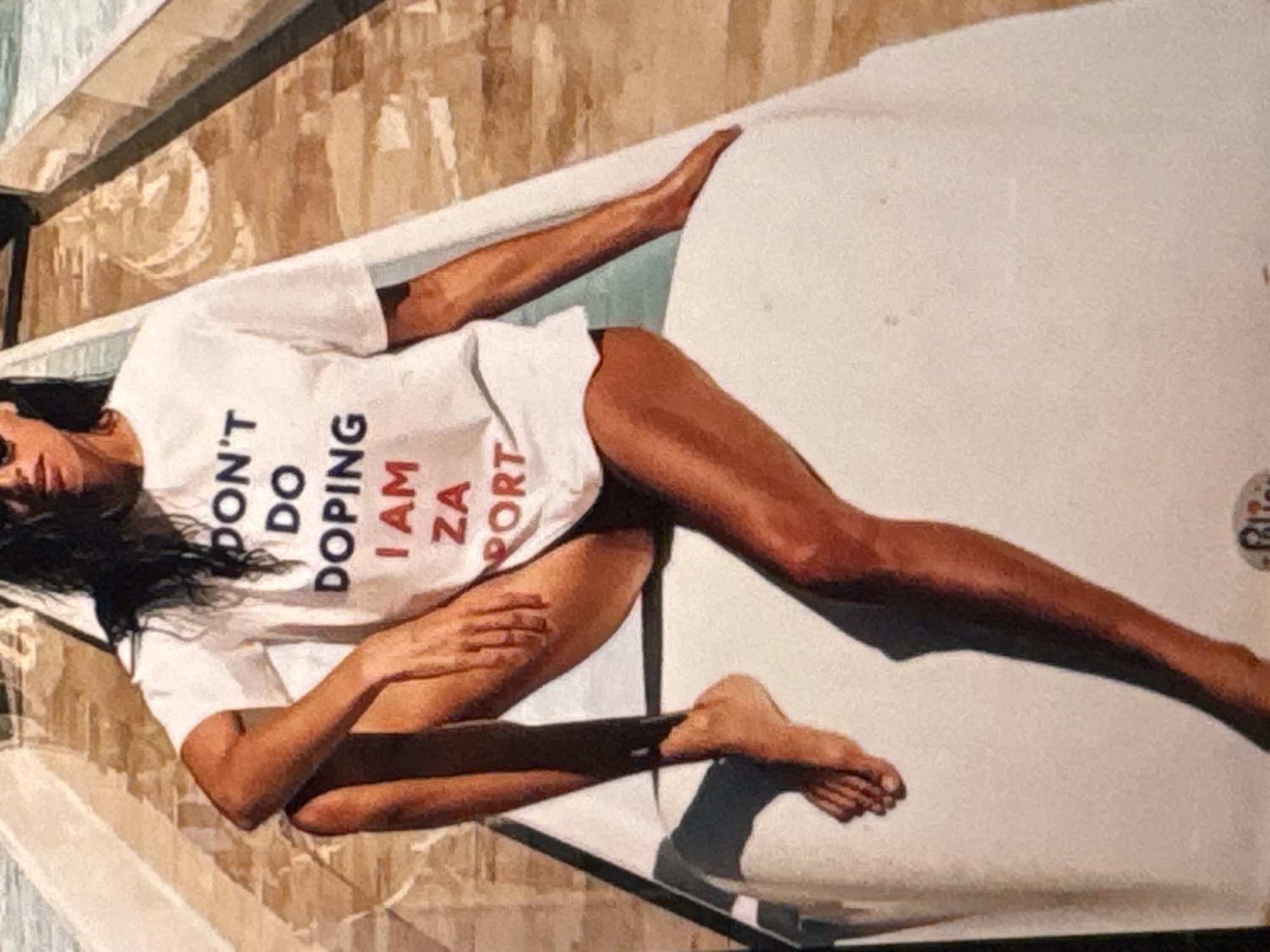 A woman with dark, wet hair and sunglasses is sitting on a white surface, possibly a boat or a lounger, outdoors. She is wearing a white t-shirt with text printed in dark blue and red letters. The text reads: "DON'T DO DOPING I AM ZA SPORT". Her legs are crossed, and her bare feet are visible. The background consists of a textured surface with diagonal lines, suggesting a walkway or a textured deck. The overall lighting is bright, indicating it is daytime. The image does not provide any explicit indicators for the location in Kuta Selatan, Indonesia.