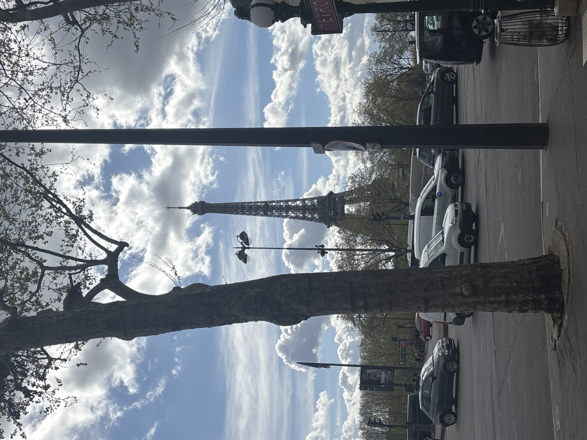 The image depicts a daytime urban street scene in Paris, France. The Eiffel Tower is prominently featured in the central background, a dark metal lattice structure against a partially cloudy sky with blue patches and white cumulus clouds.
In the immediate foreground on the left, a large, dark-barked tree trunk with sparse branches and nascent foliage rises vertically, partially obscuring the left side of the scene. On the right, a slender black utility pole extends vertically, with a black wire-mesh trash bin at its base. A traditional black "METRO" sign with red lettering is affixed to a decorative street lamp pole on the far right.
The mid-ground displays a multi-lane paved road with several vehicles. A dark grey Fiat 500 hatchback is positioned in the left lane, angled as if turning left. Behind the foreground tree, a white panel van is partially visible. Further right, a dark-colored SUV or wagon is present in traffic. In the distance, a tall black street lamp pole with two luminaires is visible to the right of the central tree. To the left of the Eiffel Tower, a dark flag on a flagpole stands next to a billboard displaying text that includes "RAMA." A line of trees with sparse light green foliage partially obscures the base of the Eiffel Tower and the far side of the street, where indistinct human figures and additional vehicles are present near traffic signals.