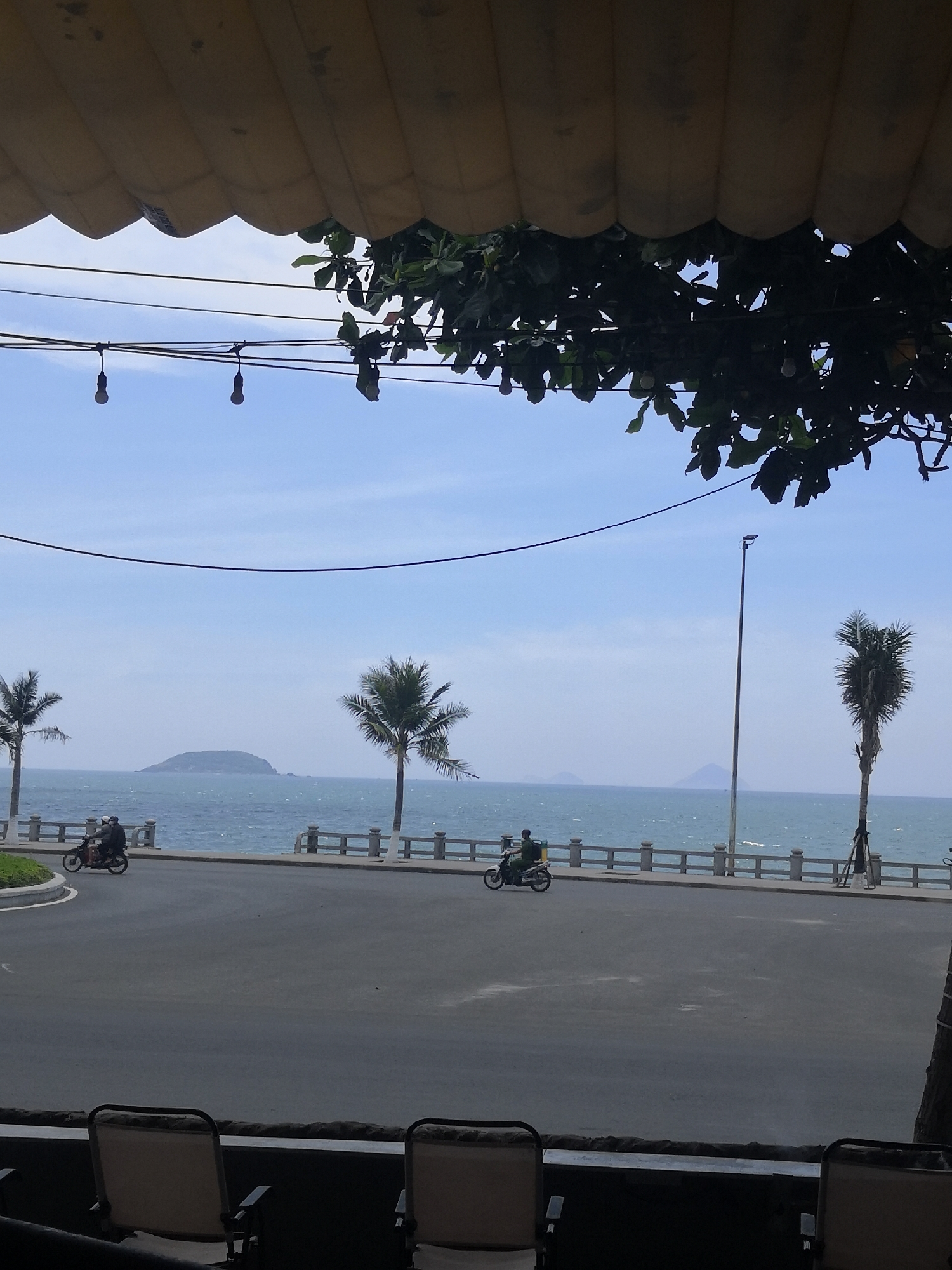 This is a photograph taken in Bac Nha Trang Ward, Vietnam, offering a view of the ocean from beneath a yellow awning and a tree canopy. The foreground is partially obscured by dark shapes, possibly furniture or railings, with two chairs visible. Beyond this, a paved road runs parallel to the sea, with a white railing separating it from the water. Several palm trees are scattered along the coastline. On the road, two motorcycles with riders are visible, one closer to the left and the other further to the right. The ocean stretches out to the horizon, with a small island visible in the distance. A tall, slender lamppost stands near the right side of the frame. The sky is a light blue with some scattered clouds. The overall scene depicts a coastal road on a clear day.