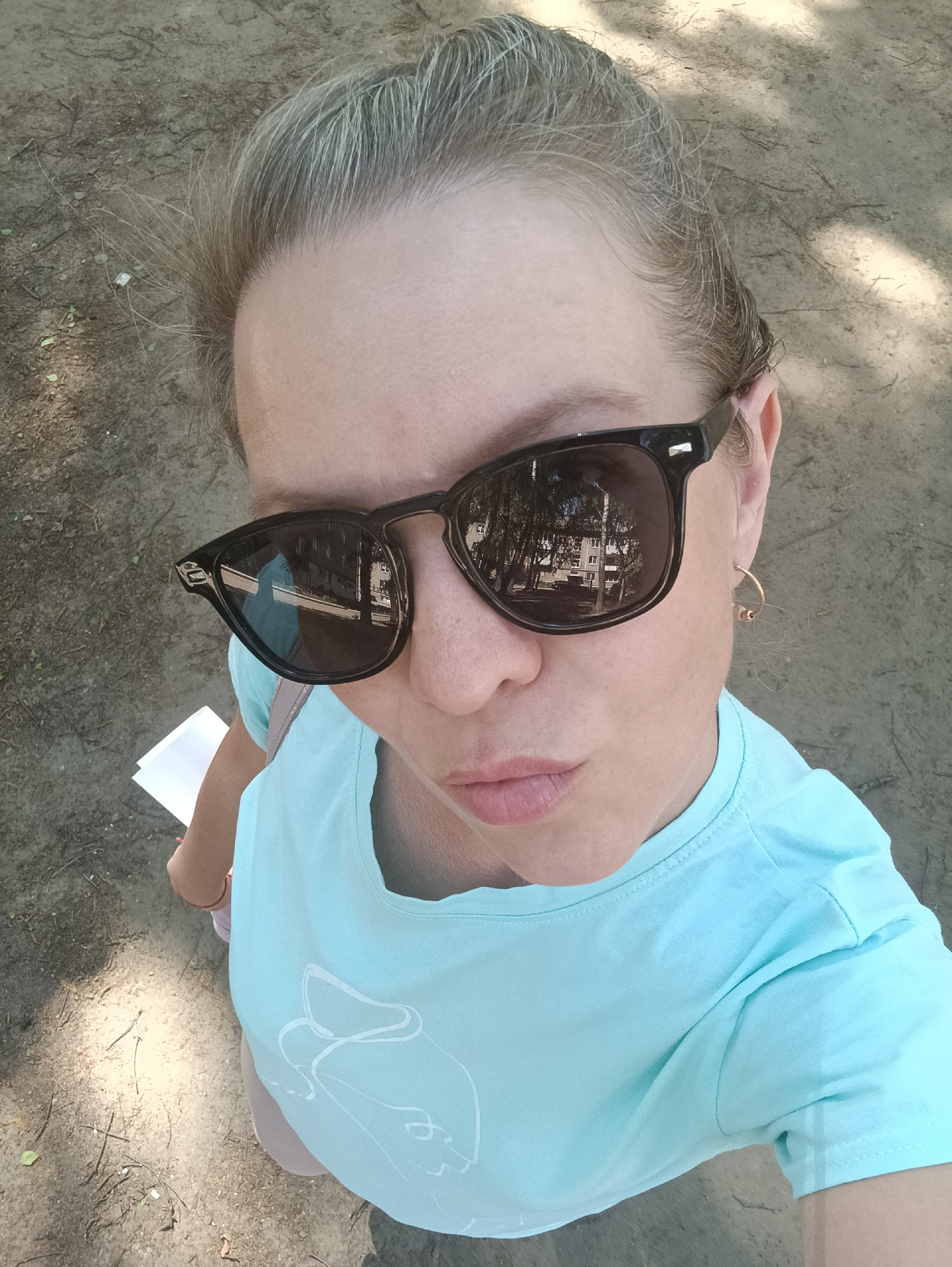A woman with fair skin and blonde hair pulled back is taking a selfie from a high angle. She is wearing dark sunglasses that reflect a building with multiple windows and surrounding trees. She also wears a light blue t-shirt and a gold hoop earring. In her hand, she holds a white paper. The ground is visible in the background and appears to be an outdoor space covered in dirt and small twigs. Based on the reflection in the sunglasses, the location is Chaikovsky, Russia, and the reflection shows a typical Russian urban landscape.