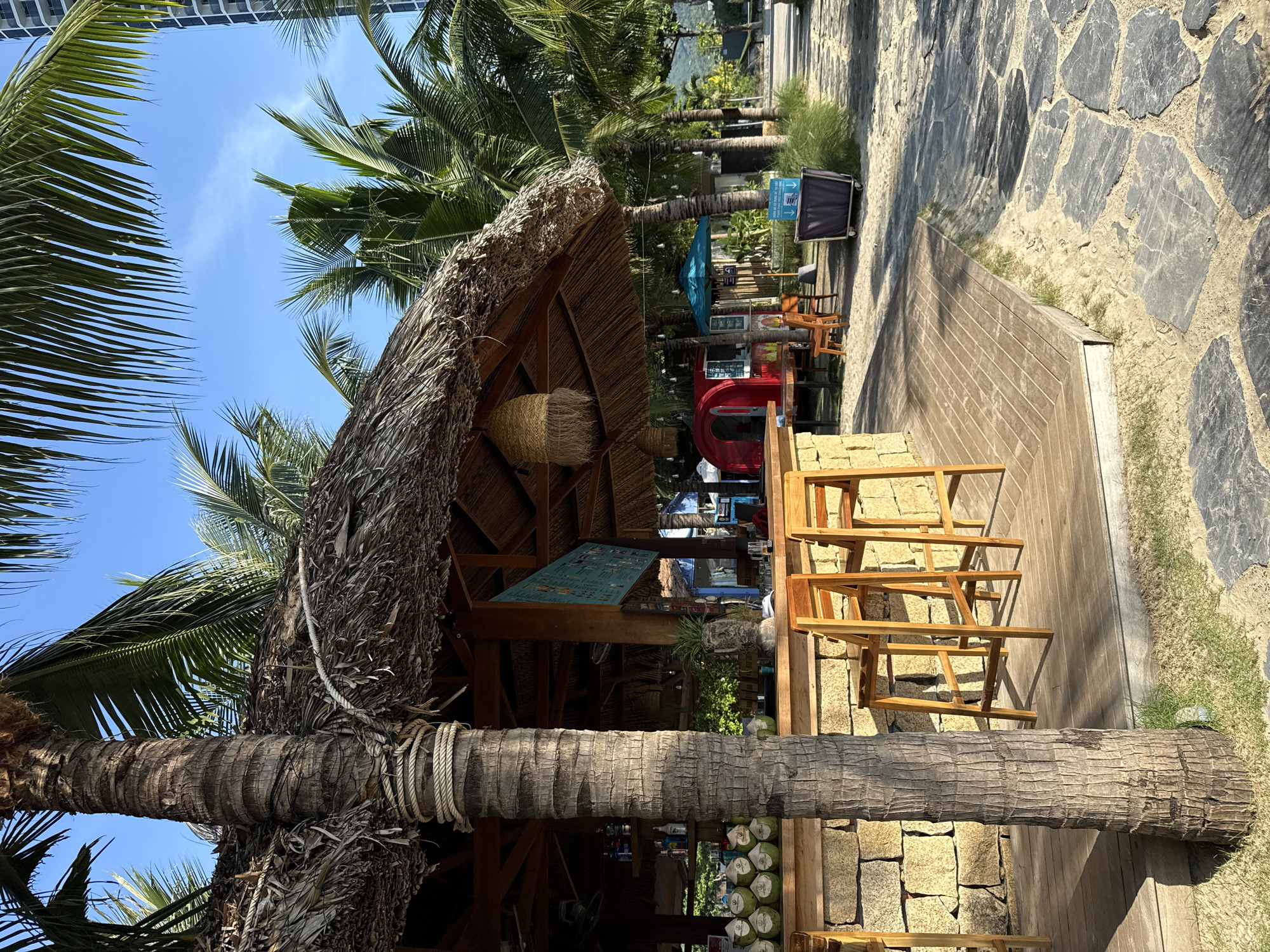 This image depicts an outdoor bar area on a sunny day, likely in a tropical beach setting, given the palm trees and thatched roof. The bar itself is constructed from stone blocks and a wooden countertop. Two wooden bar stools are placed in front of the bar. To the left of the bar, a stack of coconuts is visible. In the background, there are tables and chairs under umbrellas, a red food stall, and a tall modern building. A paved walkway made of irregular stone slabs borders the sandy area. The overall scene suggests a relaxed, vacation-like atmosphere. The location is specified as Bac Nha Trang Ward, Vietnam.