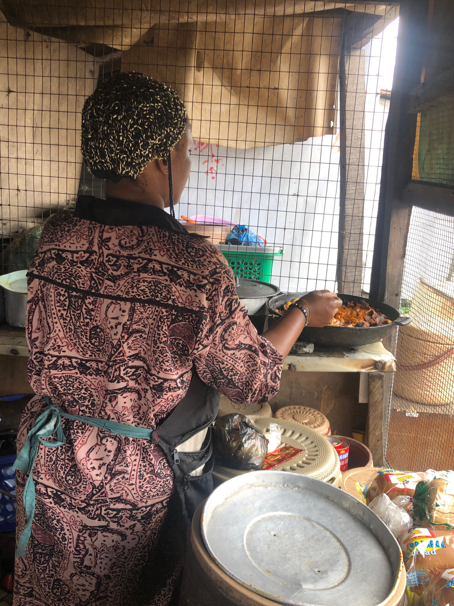 The image depicts a street-side food stall in Lagos, Nigeria. A woman, seen from behind and from the waist down, is preparing food in a large, dark-colored pan over an apparent heat source. She is wearing a patterned shirt with black and pink paisley designs and a teal apron tied around her waist. Her hair is styled in a bun with visible braids. The stall is constructed with a wire mesh enclosure and wooden beams, with a tan cloth draped behind it. In the foreground, various food items and containers are visible, including packaged bread, a large metal lid, and a plastic basket. The overall environment suggests a bustling urban setting.