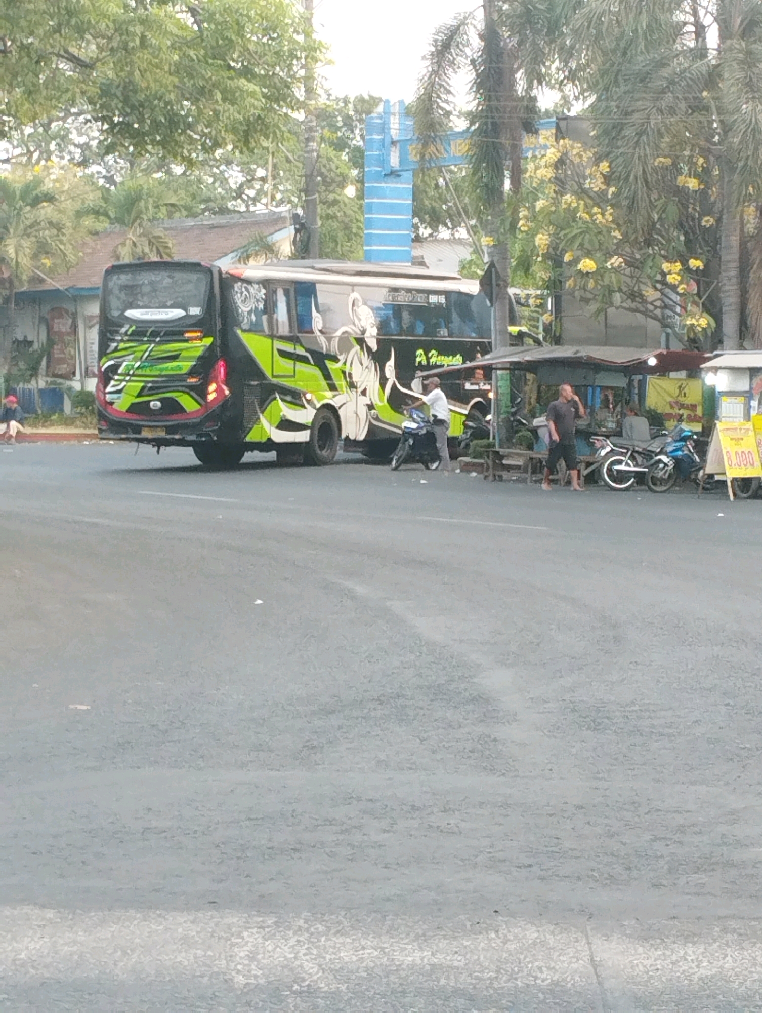 The image shows a street scene in Kecamatan Pati, Indonesia, with a large tour bus parked on the right side of the road. The bus is predominantly black and green with decorative graphics. In front of the bus, there is a small outdoor food stall with several people standing around. One person is walking away from the stall, while another is sitting at the stall. Two motorcycles are parked near the stall, and one person is standing next to a motorcycle. The road is paved, and there are trees and buildings visible in the background. The sky is overcast.