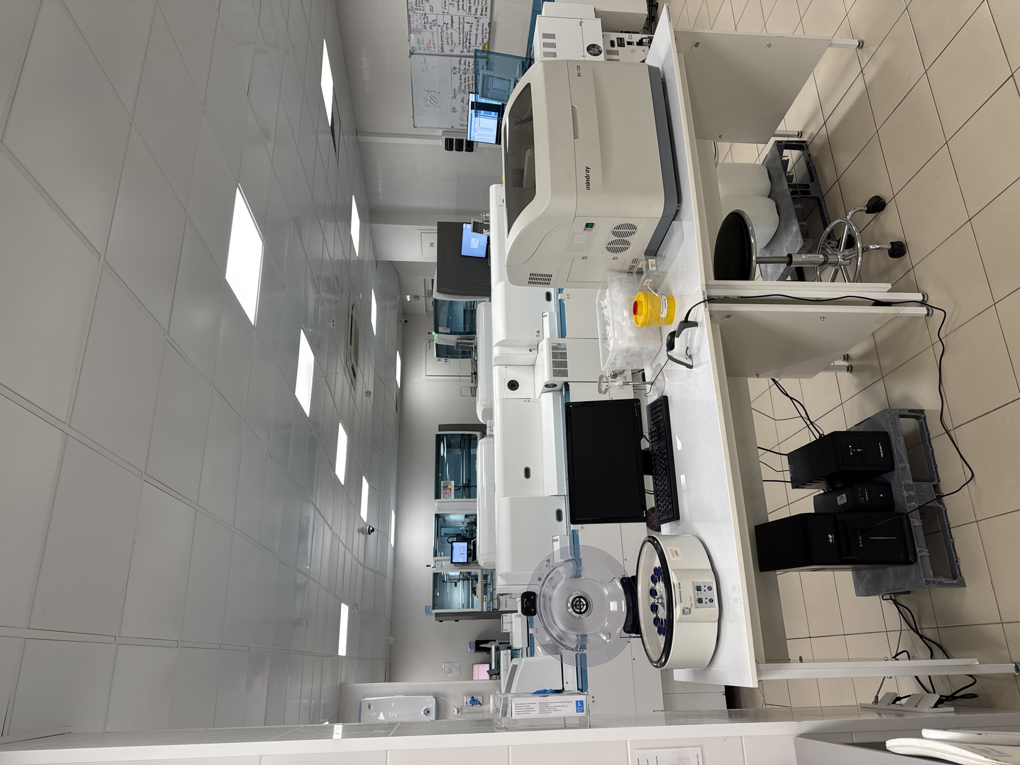 This image shows a laboratory setting with various scientific equipment. The room is well-lit with fluorescent ceiling lights and has tiled flooring. In the foreground, a white lab bench is set up with a computer monitor and keyboard, a centrifuge with blue tubes, and a larger analytical instrument labeled "Mindray BS-120". Underneath the bench, there are several black computer towers and uninterruptible power supplies. In the background, more laboratory equipment is visible, including automated analyzers and other large machines. There are also whiteboards on the wall with handwritten notes and diagrams. There are no people visible in the image. The environment suggests a clinical or research laboratory, likely in a healthcare facility or research institution. The presence of specific scientific equipment such as centrifuges and automated analyzers points to a focus on laboratory diagnostics or biomedical research. There is no information in the image to suggest a specific location in Moscow, Russia.