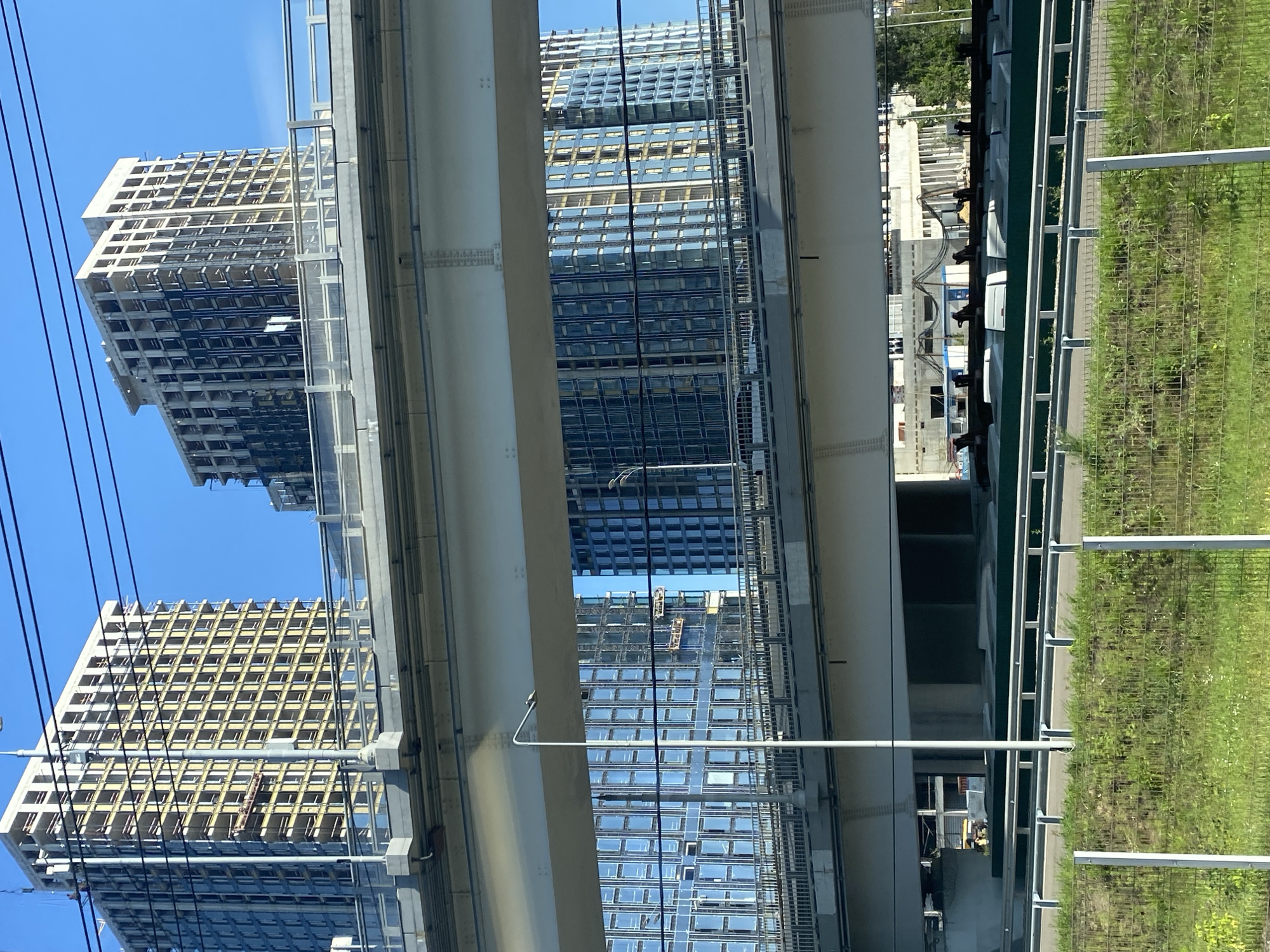 In the city of Unknown, Unknown, a cityscape is depicted under a clear blue sky. The foreground features a grassy area with a chain-link fence, beyond which a multi-lane road is visible with guardrails. Elevated roadways, supported by concrete and steel structures, dominate the mid-ground. Through the gaps in these elevated structures, several tall buildings are visible. Two buildings on the left appear to be under construction, with their structural frames exposed and scaffolding present. One of these buildings has a façade of yellow square panels. To the right, two completed buildings are seen with reflective glass facades. The sky is a vibrant blue, with a few overhead power lines crossing the frame. The lighting suggests it is daytime, likely late morning or early afternoon, given the bright illumination and shadows.