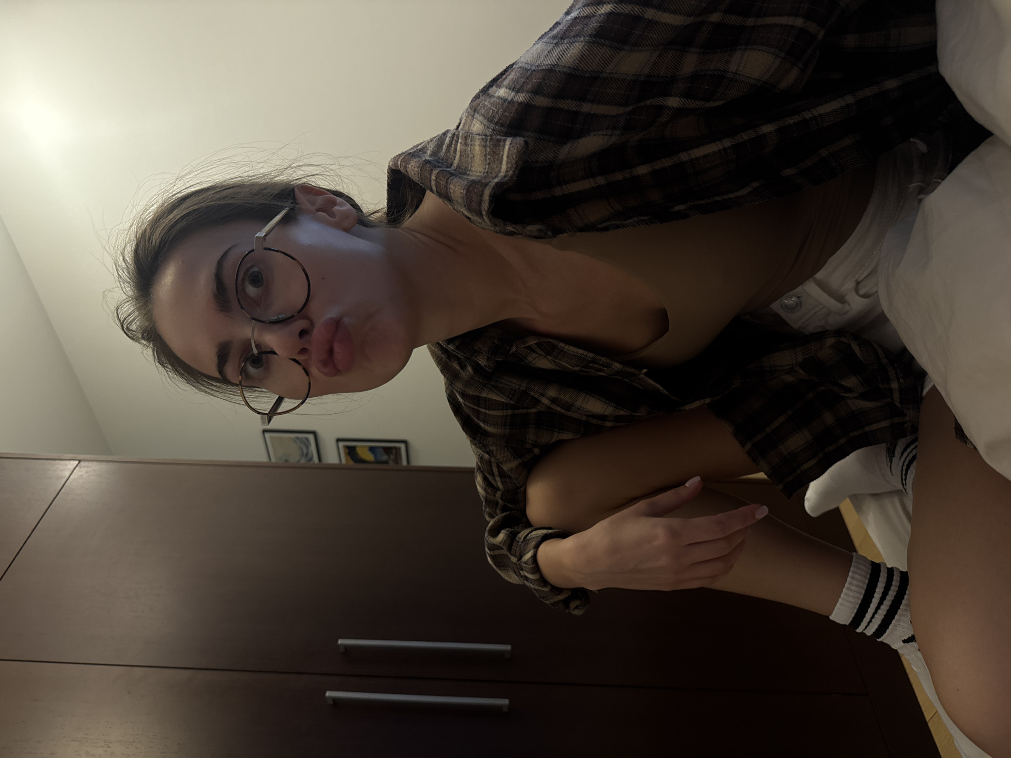 A medium shot of a woman in an indoor setting, with the location being the city of Unknown, Unknown. The woman is wearing round glasses, a brown plaid shirt, and a tan top. She has her legs pulled up to her chest and is wearing white socks with black stripes. The background includes a dark brown wardrobe with two silver handles and three framed pictures hanging on the wall behind the wardrobe. A white pillow is also visible in the scene.