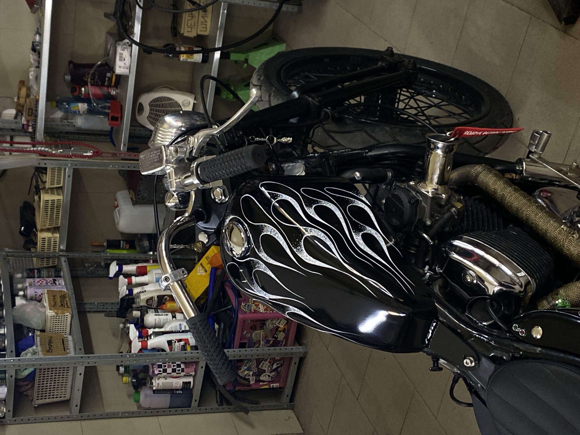 The image shows a black motorcycle parked indoors, likely in a workshop or garage. The motorcycle has a custom paint job with white flames on the fuel tank. Various shelves filled with cleaning supplies, tools, and other items are visible in the background. The scene is well-lit, and the motorcycle is the primary subject of the photograph. There are no people visible in the image. The location is identified as Agronom, Russia, but no specific details about the environment beyond it being an indoor workshop are evident.