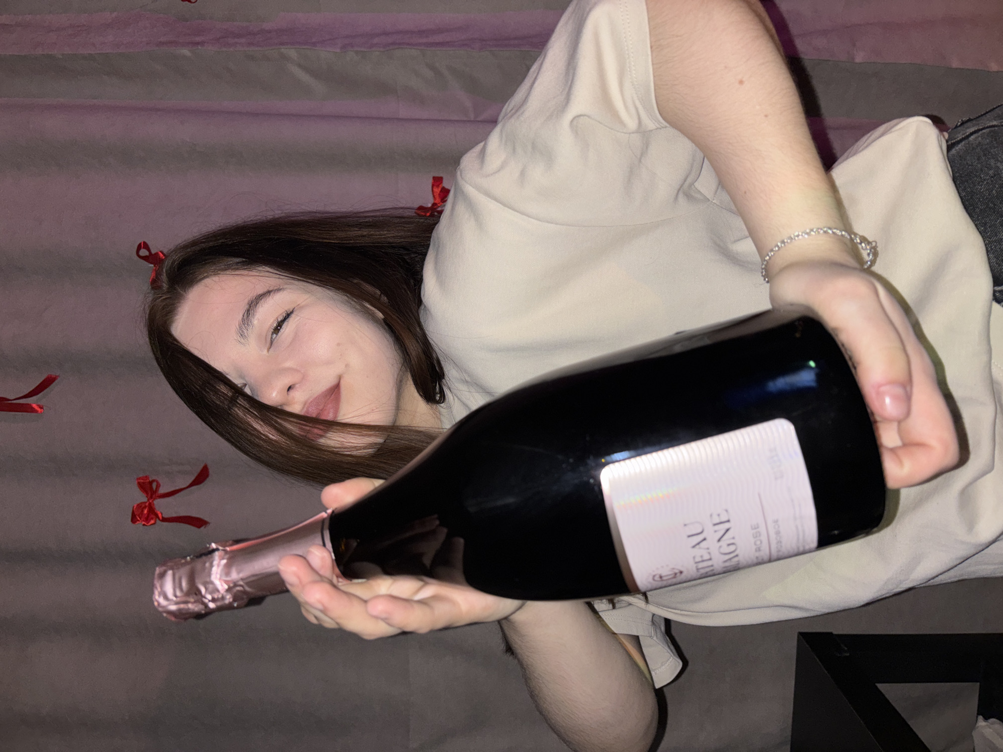A young woman with long, dark hair is captured in a close-up, high-angle shot, lying down on what appears to be a bed or sofa in Perm, Russia. She is wearing a light-colored t-shirt and has a silver-toned bracelet on her left wrist. Her eyes are partially closed, and a gentle smile plays on her lips, conveying a relaxed and happy emotion.
With both hands, she holds a dark-colored bottle, likely champagne or sparkling rosé, positioned prominently in the foreground. The bottle features a pink foil cap and a white label, on which partial text is visible, including "ATEAU," "PAGNE," and "ROSE." Scattered around her on the soft, gray-purple fabric surface are several small red ribbon bows, hinting at a festive, romantic, or celebratory occasion. The lighting suggests an indoor setting, and the overall mood is intimate and cheerful.