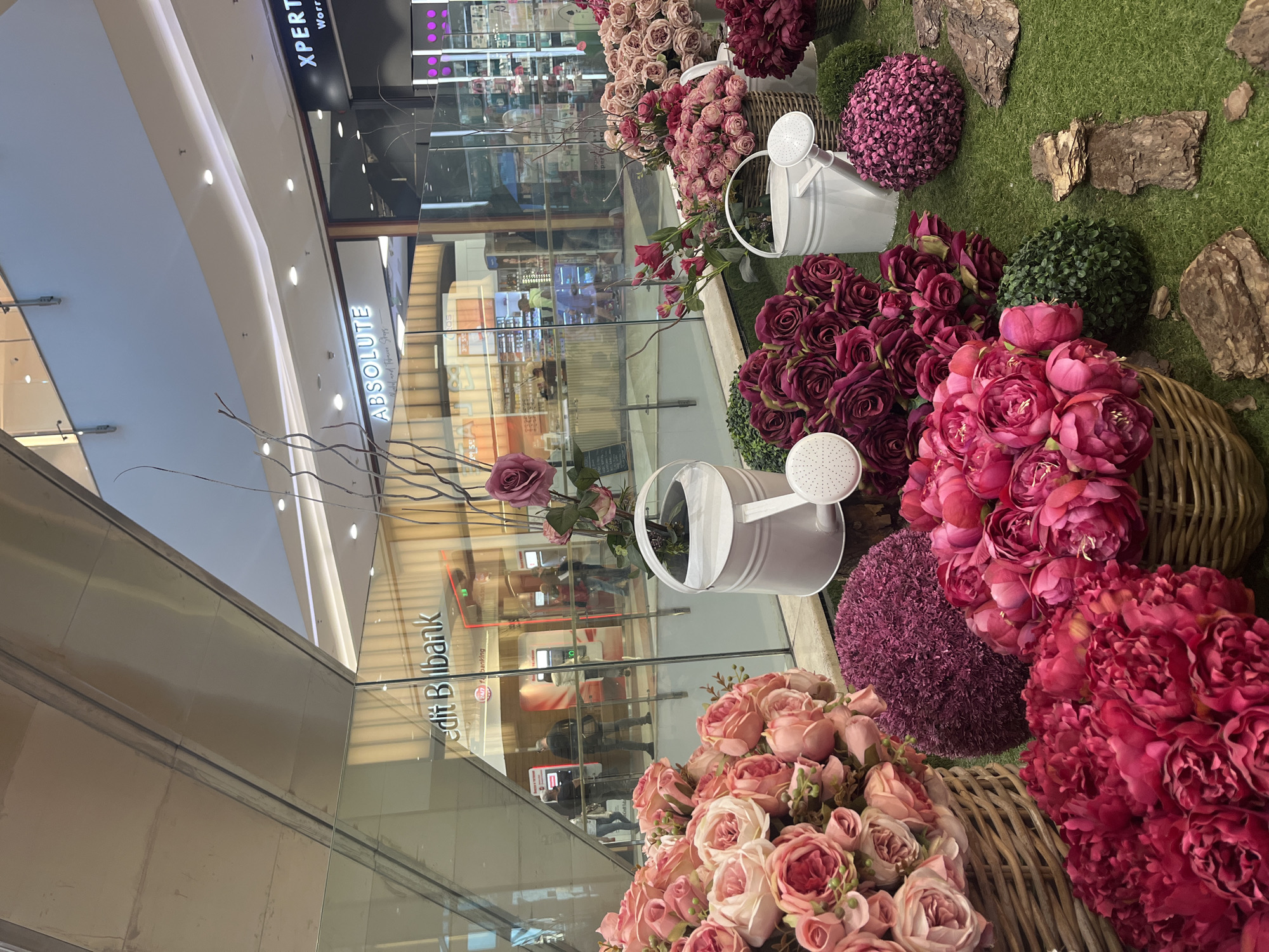 The image shows a floral display in a shopping mall in Sofia, Bulgaria. The display consists of various artificial flowers, including roses and peonies, arranged in baskets and watering cans on a patch of artificial grass. The flowers are primarily pink and purple in color. Pieces of tree bark are also incorporated into the display. The display is positioned in front of glass storefronts of several businesses, including "Credit Bilbank" and "Absolute." The reflections in the glass storefronts show people walking inside the stores. The mall has a brightly lit interior with a multi-level design. The "XPERT World" signage is visible on the top floor.