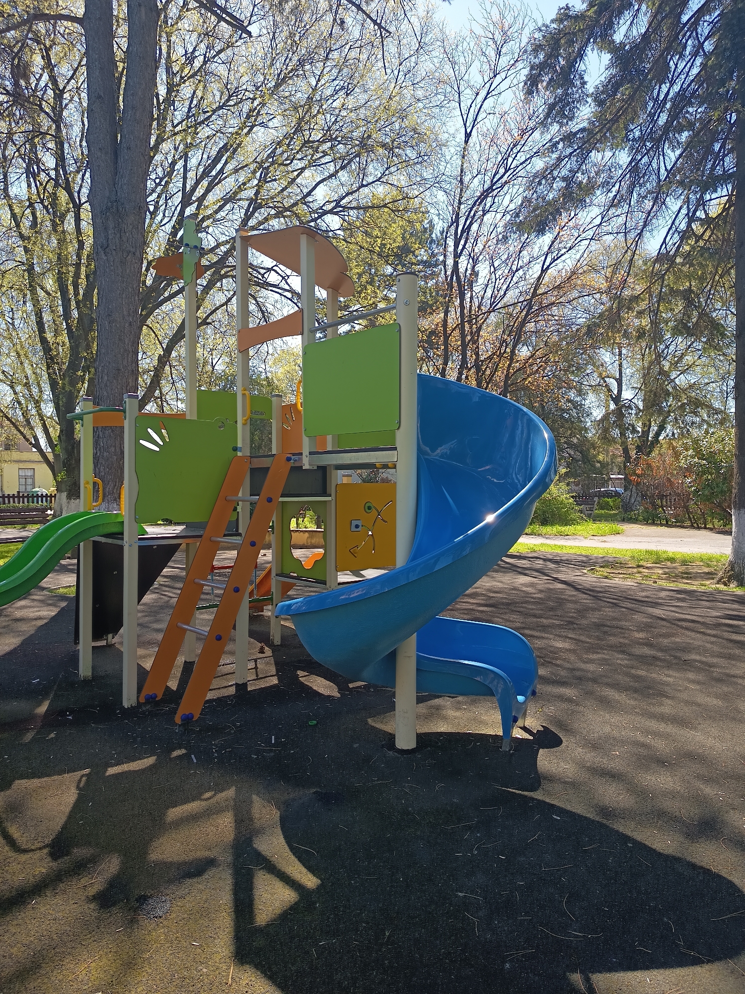 The image shows a brightly colored playground structure in what appears to be a park or recreational area. The structure features green and orange accents, a blue slide, and a ladder for access. The ground is covered in a dark material, possibly rubberized safety surfacing. The background includes trees with sparse foliage and a building in the distance. The photograph was taken during the day, with shadows cast by the structure and trees. The location is Sungurlare, Bulgaria. There are no people visible in the frame.