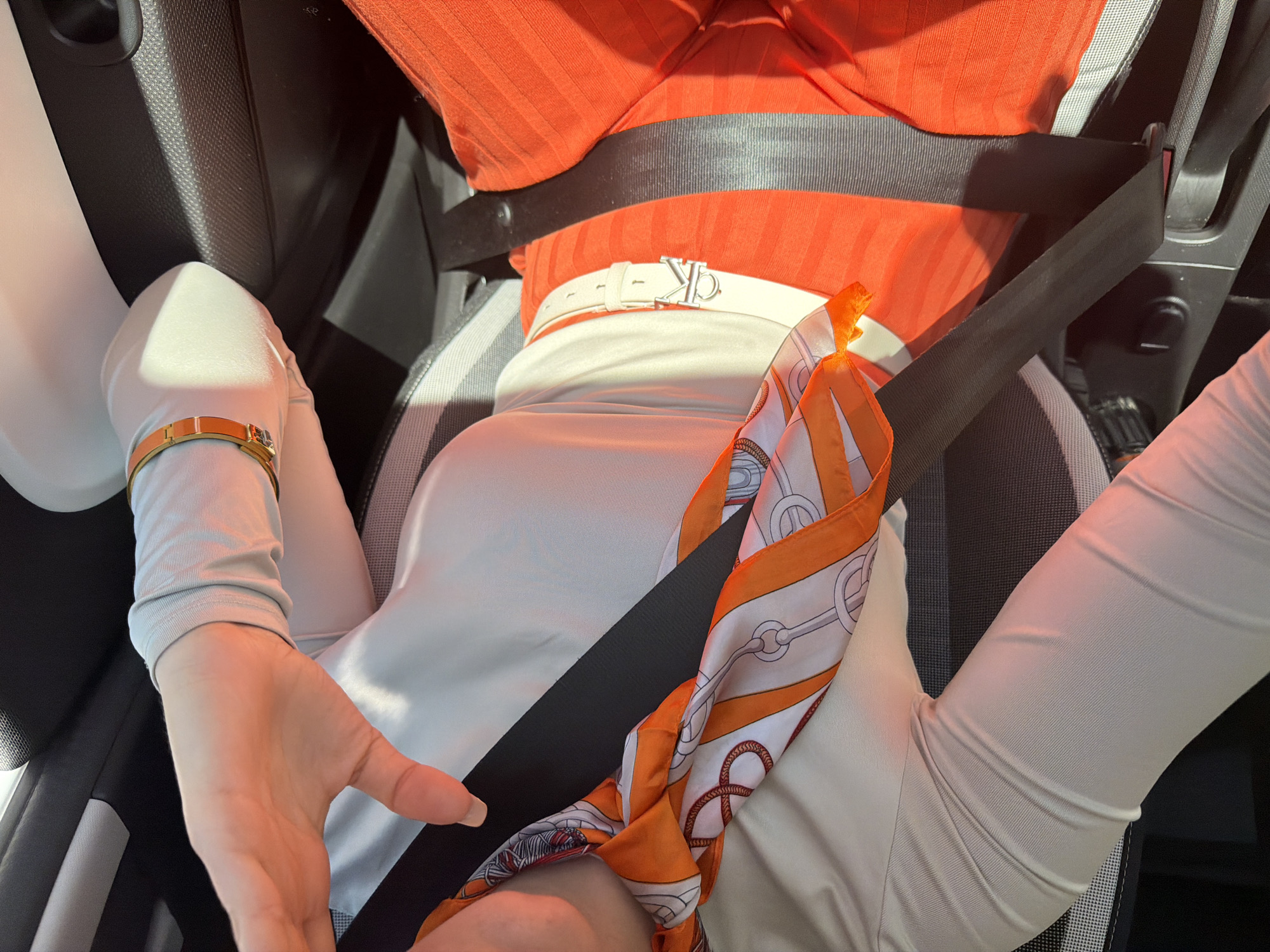 A person is shown from above, seated in a car. They are wearing an orange top, a white top, and a white belt with a "CK" buckle. They are also wearing a white top with long sleeves and a gold bracelet. An orange and white scarf is draped over the seatbelt. The environment appears to be the interior of a car, and the exact location in Sofia, Bulgaria cannot be determined from this image alone.