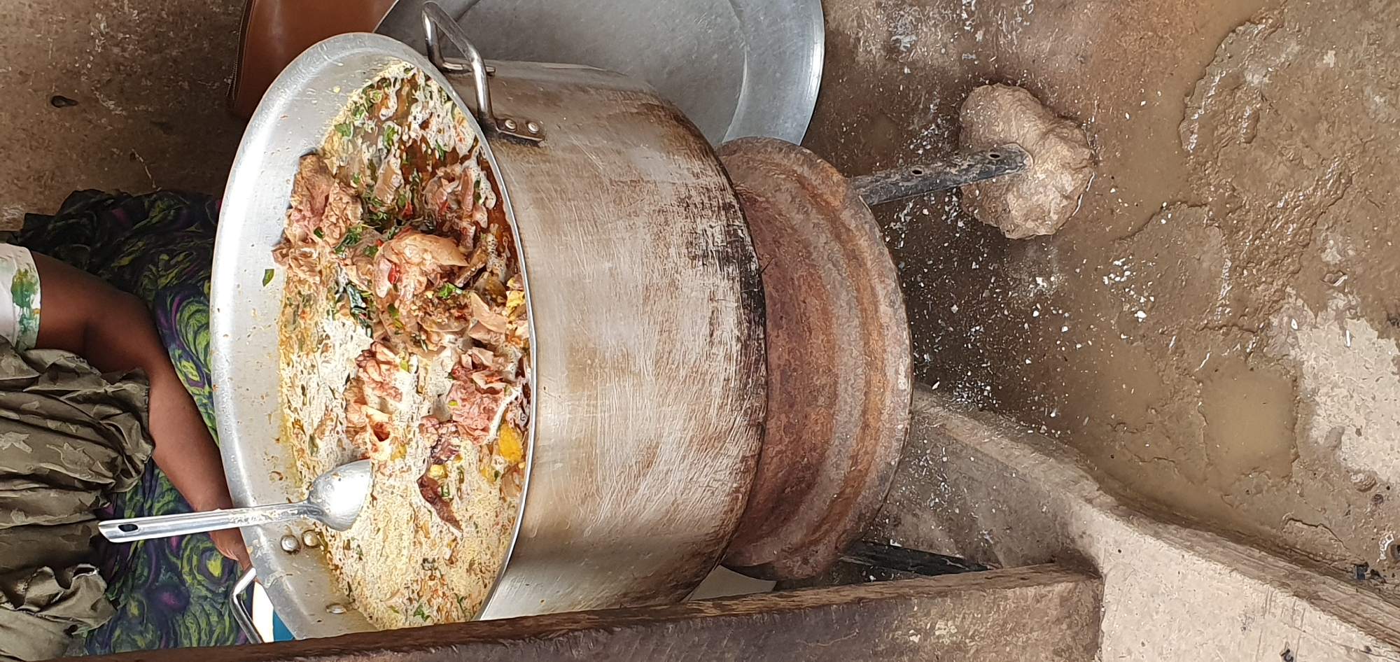 The image shows a large metal pot filled with a stew-like dish containing meat and vegetables, placed on a rustic metal stand over a fire. A ladle rests in the pot. The scene appears to be outdoors, with a person partially visible in the background wearing a patterned cloth. The ground is muddy and wet, with scattered debris. The overall impression is of an outdoor cooking setup, likely in a market or residential area. The context of Bukuru, Nigeria, suggests a community gathering or food preparation for a local event or business.