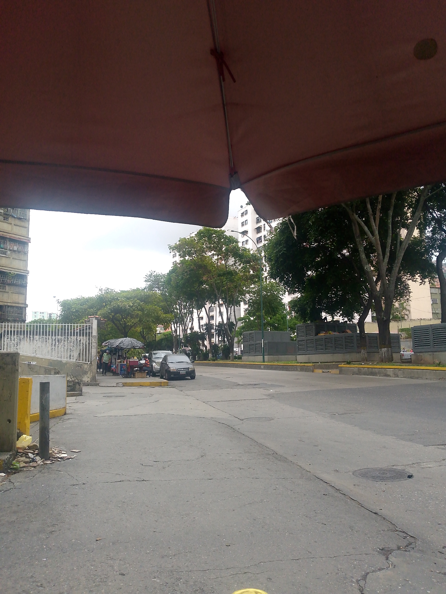 The image depicts a street scene in Caracas, Venezuela, possibly a commercial or residential area. A large brown awning dominates the foreground, partially obscuring the view. The street appears worn with cracked asphalt. To the left, there's a sidewalk area with some litter and debris near a barrier with yellow markings.
Across the street, there is a vendor operating under a black umbrella near a fence and a light gray car. Buildings are visible in the background, along with several trees. A retaining wall with horizontal louvers is visible as well. The sky is overcast. There does not appear to be any active traffic.