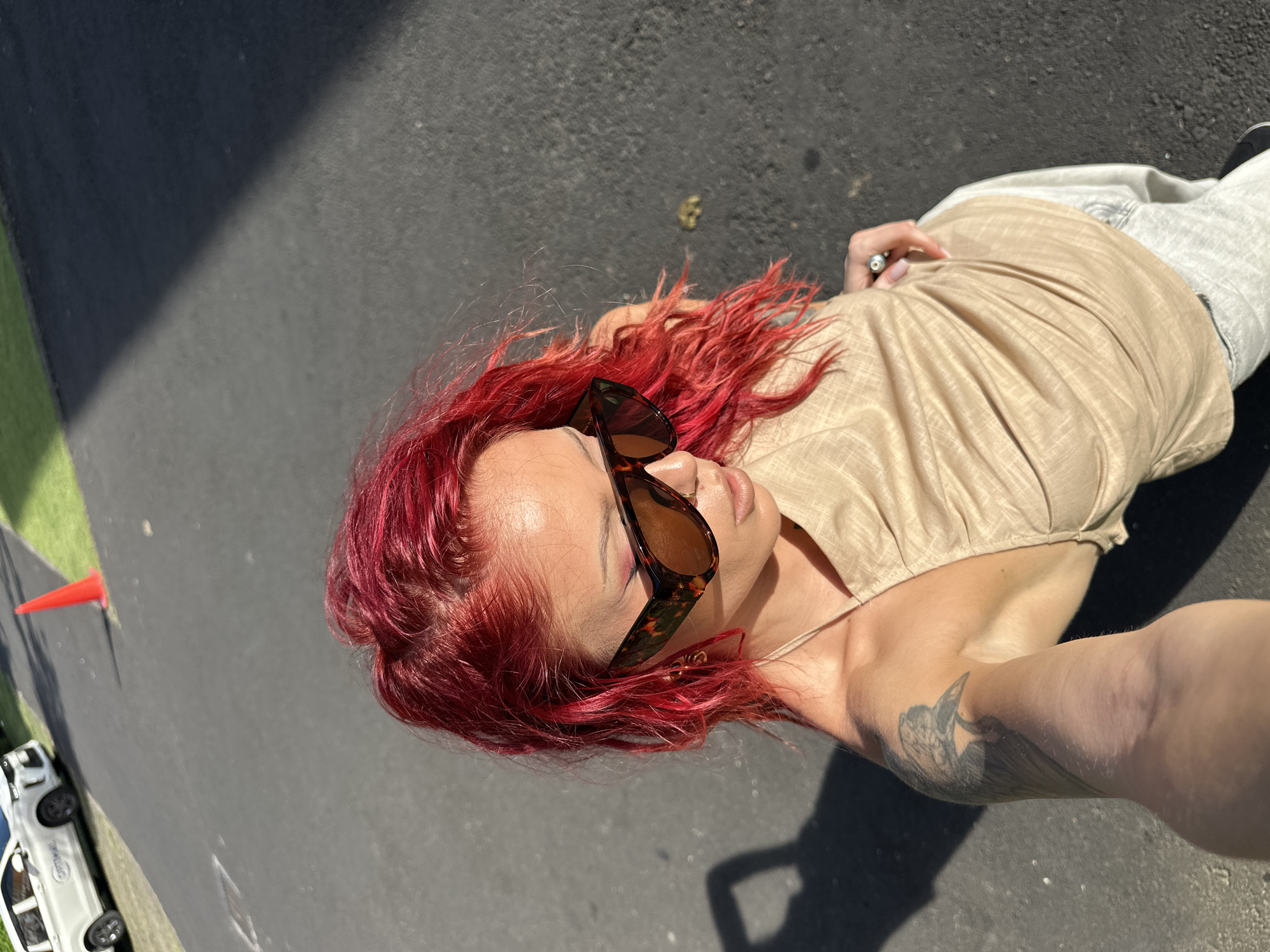 A high-angle, slightly rotated shot shows a woman with dyed red hair and sunglasses lying on an asphalt surface. She is wearing a tan top and light-colored jeans. A tattoo is visible on her right arm. In the background, a small white car is parked near a green grassy area and a red traffic cone. The location cannot be determined from this image.