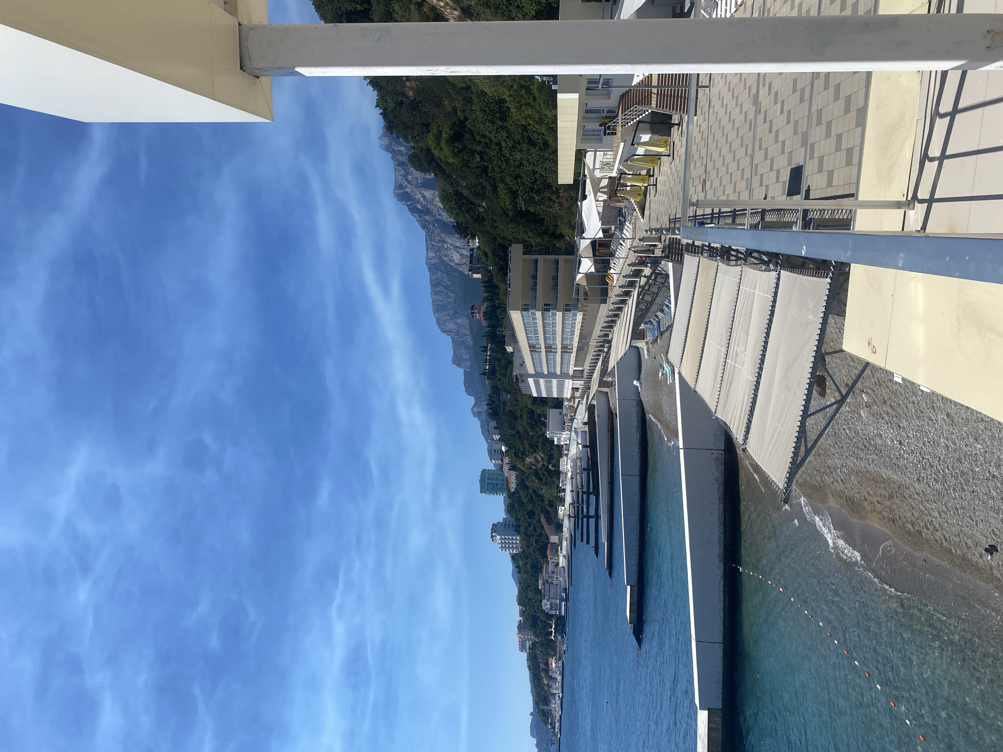 This image depicts a coastal resort area in the vicinity of Gaspry, Crimea, Ukraine. The scene features a shoreline with a pebble beach, calm blue water, and several concrete breakwaters extending into the sea. Along the beach, there are rows of white canopies providing shade over lounge chairs. A paved walkway with patterned tiles runs parallel to the beach, lined with a metal railing. In the background, a multi-story hotel with numerous windows is visible, situated on a hillside covered in lush green trees. Beyond the hotel, rugged mountains rise against a bright blue sky with scattered white clouds. A white structure with a column and railing is in the foreground, partially obscuring the view.