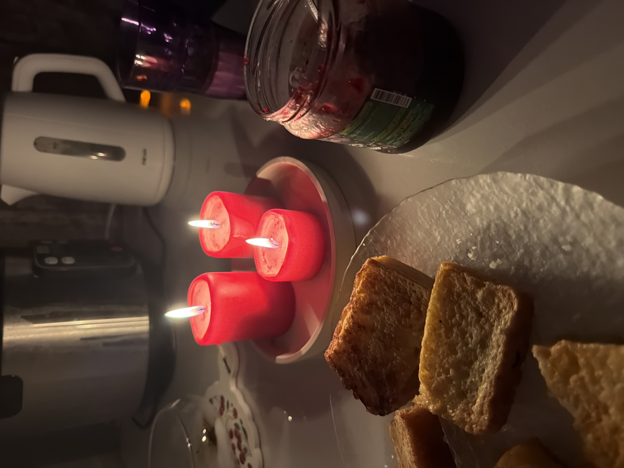 The image depicts a dimly lit indoor scene, likely in a kitchen. The focal point is a plate of golden-brown toast or fried bread slices. Beside the plate is a candle holder with three lit red candles. To the left of the candles is a white electric kettle, and further to the left, a metal pot is visible. Above the candles, a jar, seemingly containing a dark preserve or jam, is positioned. In the background, what appears to be a purple or dark-colored glass container is visible. The scene is suggestive of a cozy or intimate setting, possibly a breakfast or late-night snack setup. There is no explicit information to confirm the location in the city of Leningradskaya, Russia.