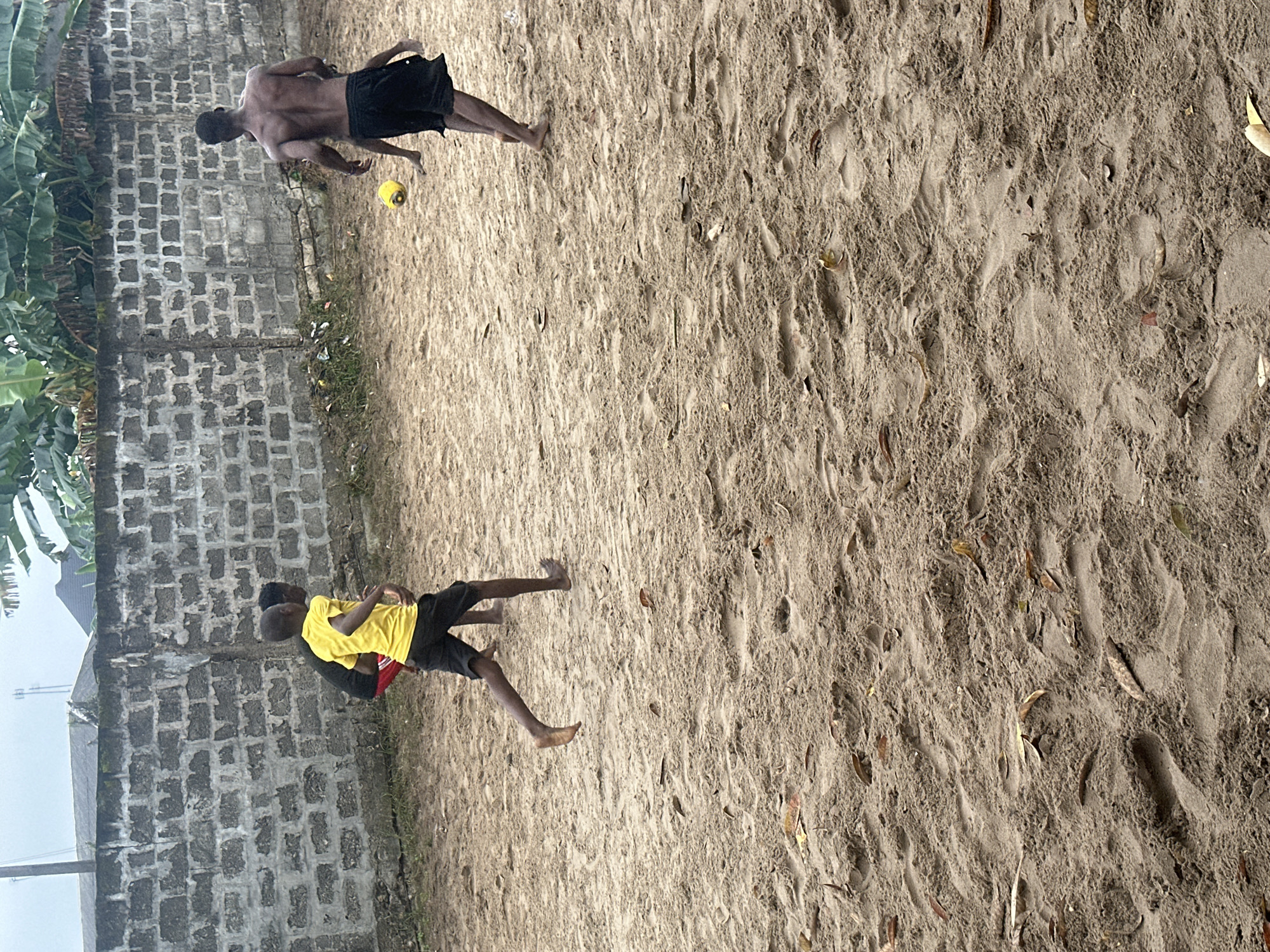 The image depicts a group of young boys playing soccer on a sandy ground in Badagry, Nigeria. In the foreground, the sandy ground shows footprints and scattered dry leaves. In the mid-ground, three boys are actively engaged in the game. One boy, wearing a yellow t-shirt and black shorts, is running, possibly towards the ball. Another boy, mostly obscured, is near him. A third, older boy, with a bare torso and black shorts, is positioned further back, looking towards the ball. A yellow soccer ball is in mid-air between the older boy and the boy in the yellow shirt. In the background, a weathered, concrete block wall serves as a boundary, with some vegetation and banana trees visible above it. The sky is overcast, suggesting a cloudy day. The scene captures a moment of casual recreation in an outdoor setting.