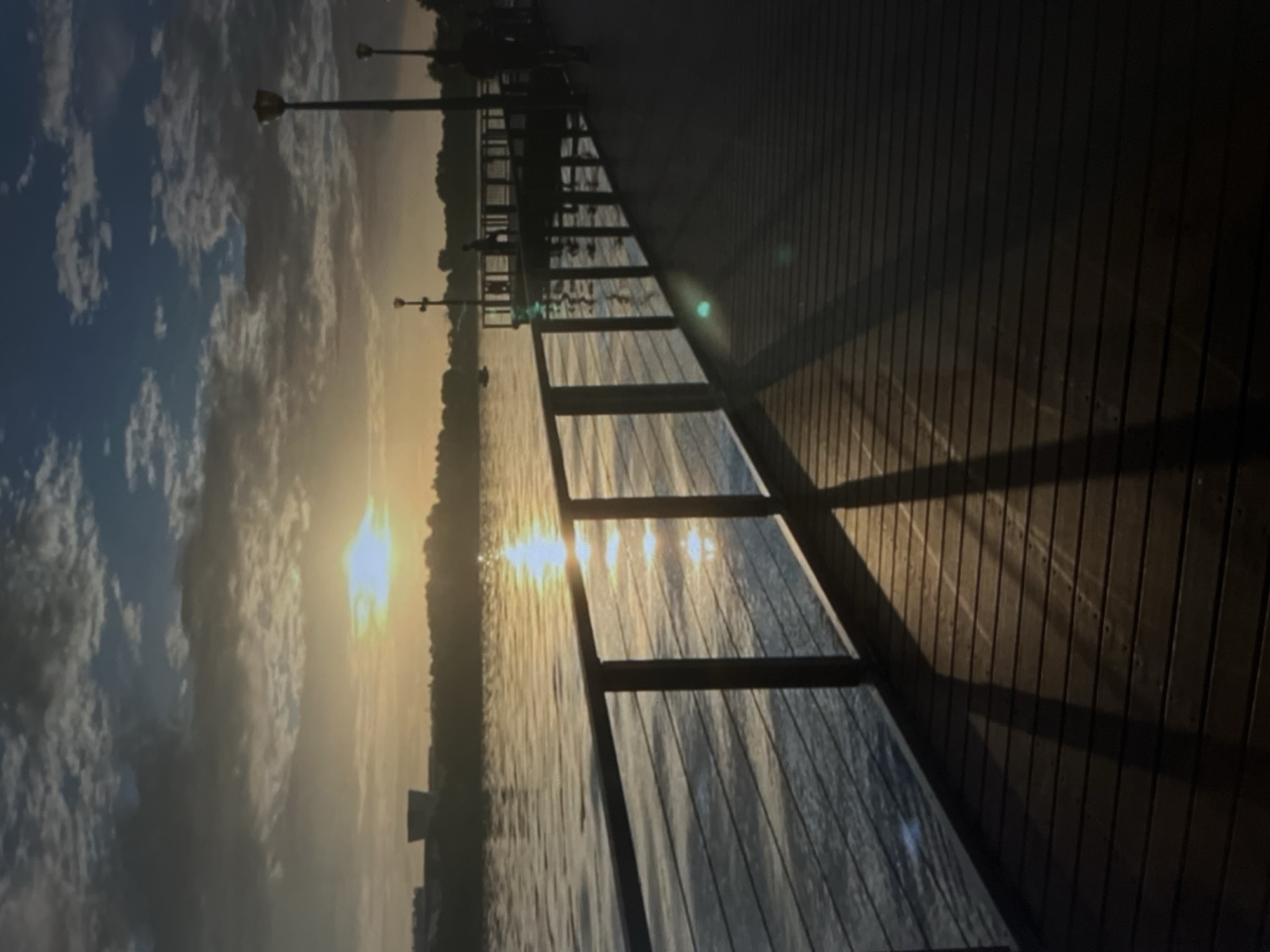 A low-angle, full shot captures a wooden pier jutting out into a body of water at sunset. The sun, a bright orb, shines through scattered clouds, casting a golden reflection on the rippling water. A metal railing with vertical supports lines the left side of the pier, separating it from the water. The wooden planks of the pier create a repeating horizontal pattern, and long shadows stretch across them from the railing. In the background, trees line the distant shore, and a cluster of buildings is visible on the left. Several lampposts stand along the pier, and a few silhouettes of people can be seen in the distance, some on a separate pier and others walking along the main pier. One person is on a boat in the water. The overall atmosphere is serene and tranquil.