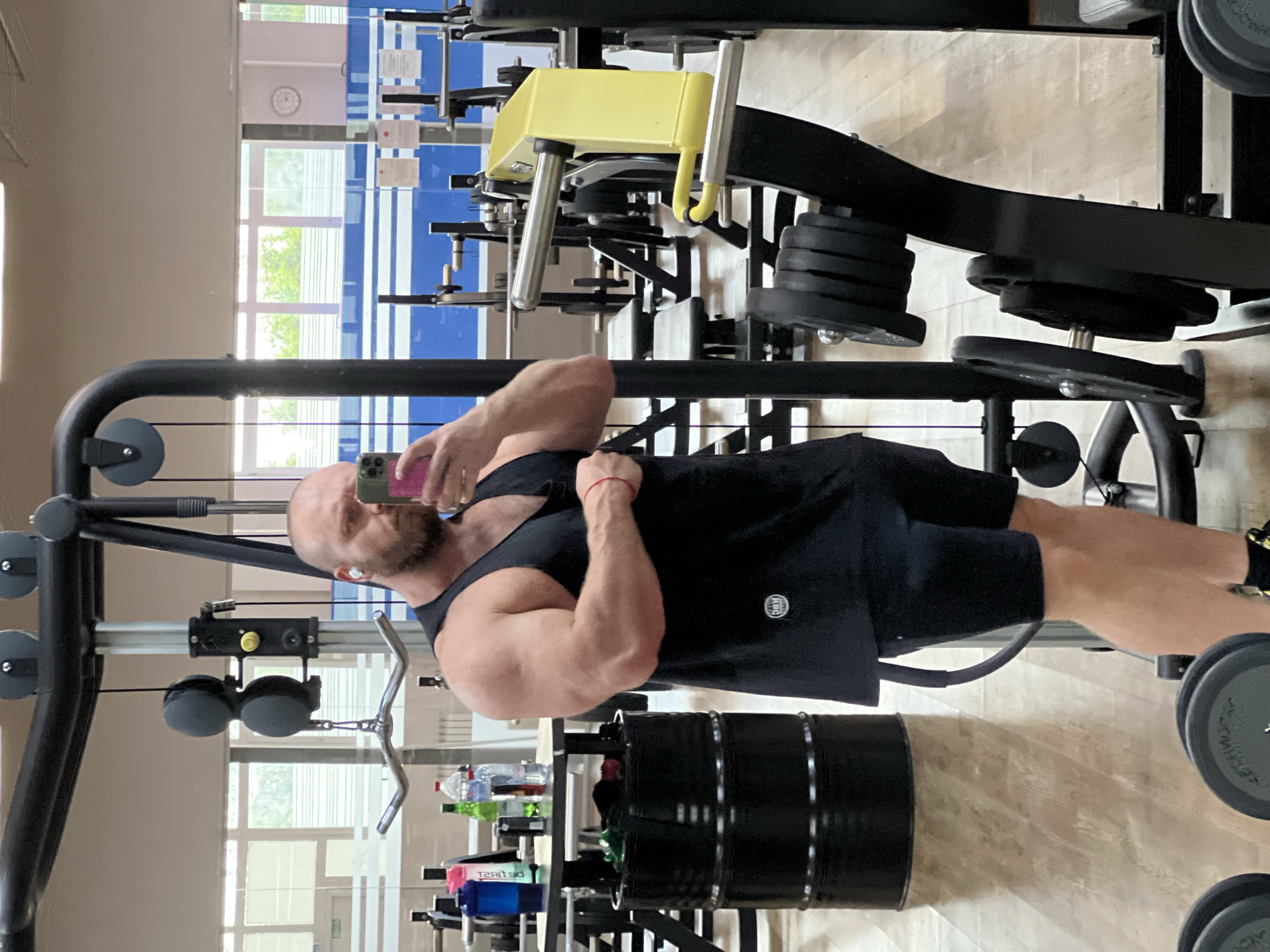 A bald, bearded man is taking a mirror selfie in a gym. He is wearing a black tank top and black shorts. He has white wireless earbuds in his ears and is holding a pink smartphone in his right hand, angled to capture his reflection. His left arm is flexed, and he is wearing a red bracelet on his left wrist. The gym appears to be well-equipped with various weight machines, including a cable crossover machine and a weight rack with stacked plates. In the background, there are windows with blinds, suggesting an indoor setting. There are also water bottles and a large black barrel visible. The floor is made of polished wood. The location is identified as a gym in the city of Chaikovsky, Russia.