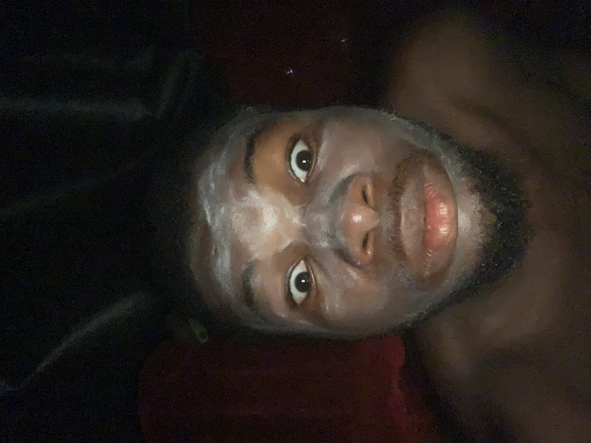 A close-up, upward-facing image shows a person with dark skin and a beard. The person has a white substance applied to their face, possibly a facial mask or cream. The person's eyes are wide open and appear to be looking directly at the viewer. The background is predominantly dark, with some reddish hues visible beneath the person's head and dark shapes suggestive of fabric or other soft materials surrounding them. There is no evidence to indicate that this image was taken in Onitsha, Nigeria, or any other specific location. No specific actions or interactions are apparent, and there are no additional identifiable objects in the scene.