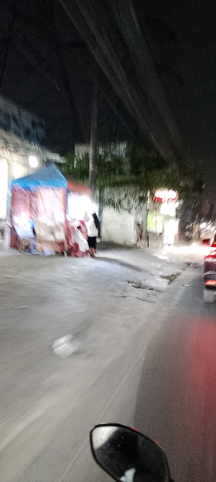 This is a night-time, motion-blurred image captured from a moving vehicle, likely a motorcycle given the presence of a mirror in the foreground. The scene appears to be a street in Satriajaya, Indonesia. On the left side of the image, there's a stall with a blue and red striped awning, selling goods. Two individuals, one wearing a dark headscarf and white top and the other in a white garment, are visible near the stall. Further down the street, a white building with some greenery around it is visible, and to the right, the back of a red vehicle is illuminated by streetlights. The road surface shows signs of damage and puddles, indicating it might have rained recently. The overall impression is of a bustling street scene at night.