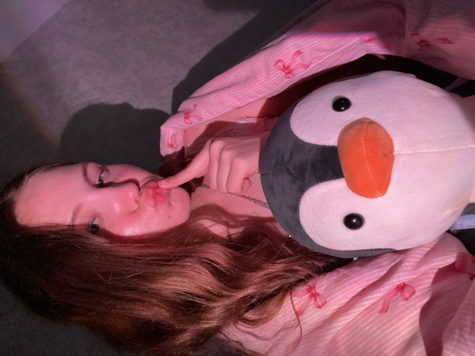 A young woman with long, wavy brown hair is holding a stuffed penguin. She is wearing a pink and white striped robe with small red bows on it. The woman is making a kissing face with her finger on her lips. The image appears to be taken indoors, possibly in a bedroom, with soft lighting casting a pink hue. There is a shadow on the wall behind her. The location is not specified in the image, and therefore cannot be determined to be in Izhevsk, Russia.